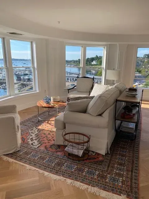 Bright living room with large bay windows, a sofa, armchair, and patterned rug overlooking a waterfront view.