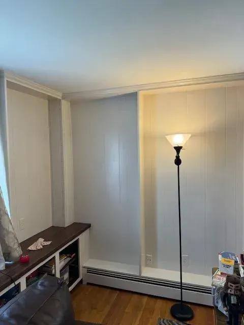 Empty living room corner with a floor lamp, white walls, baseboard heater, and a small shelf.
