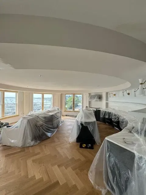 Modern living room with covered furniture, herringbone wood floor, and curved ceiling with large windows