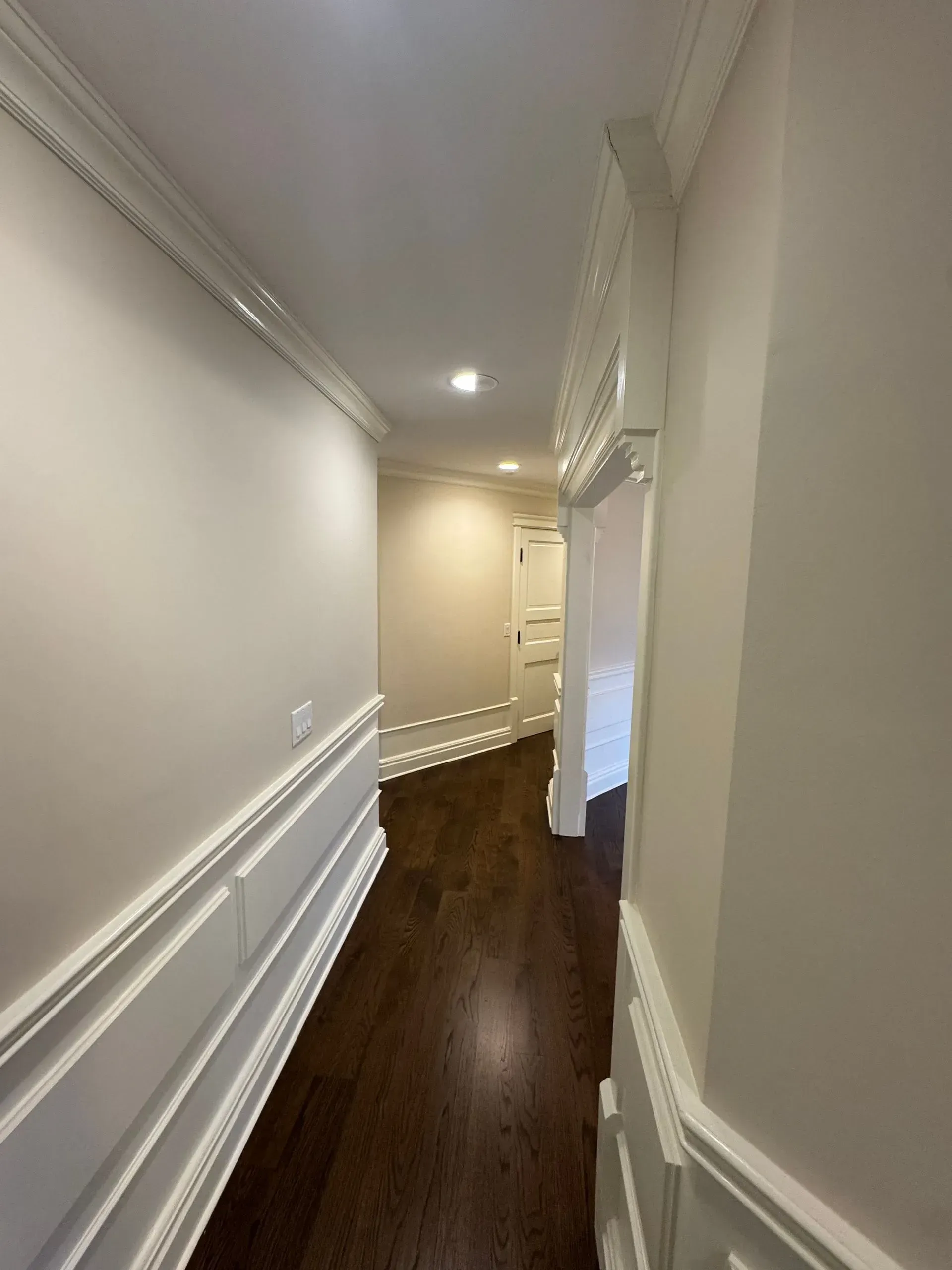 Narrow hallway with white wainscoting, beige walls, dark wood floor, and recessed ceiling lights.