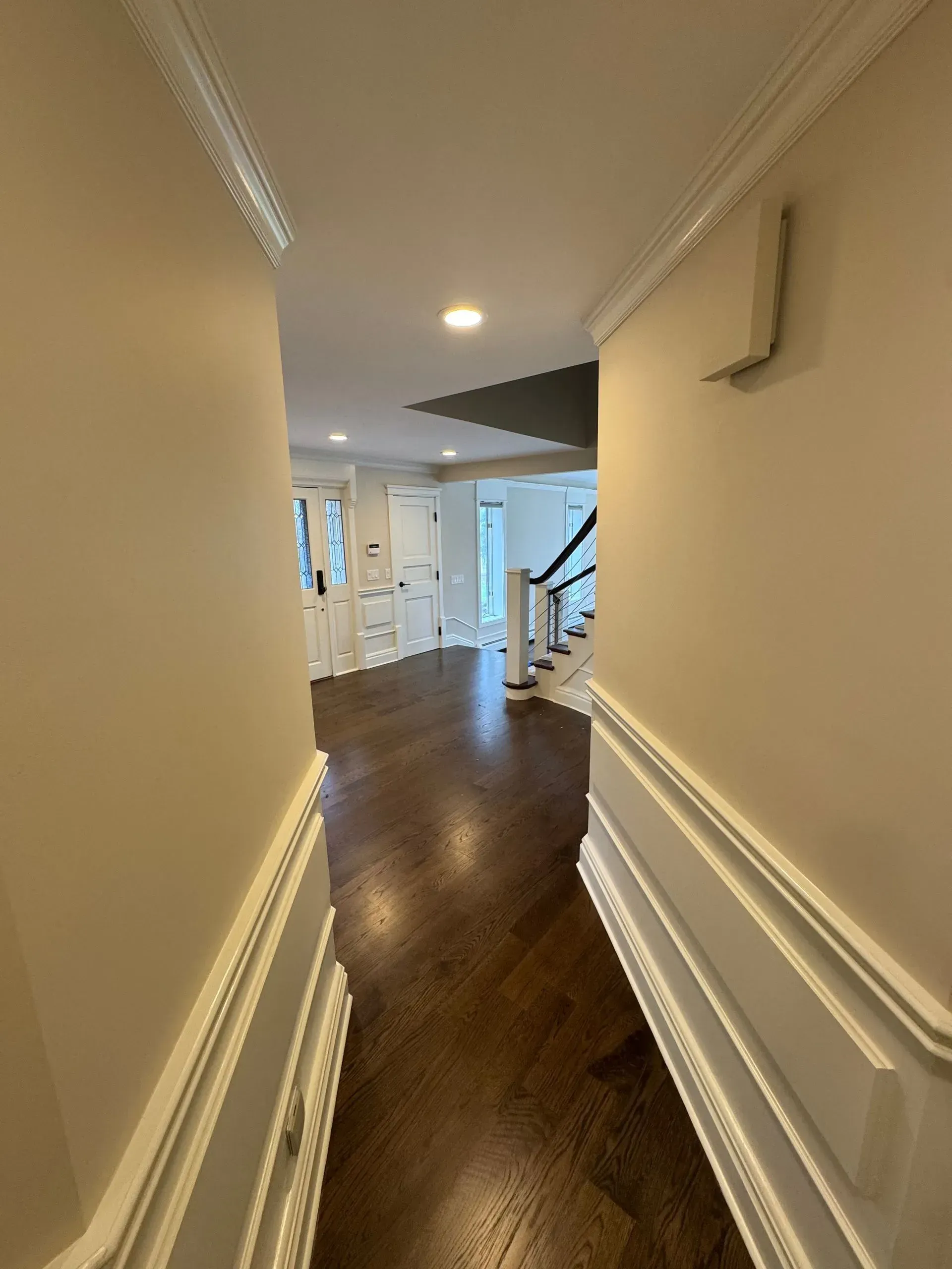 Narrow hallway with dark wood floor, cream walls, and staircase railing leading into a bright home interior