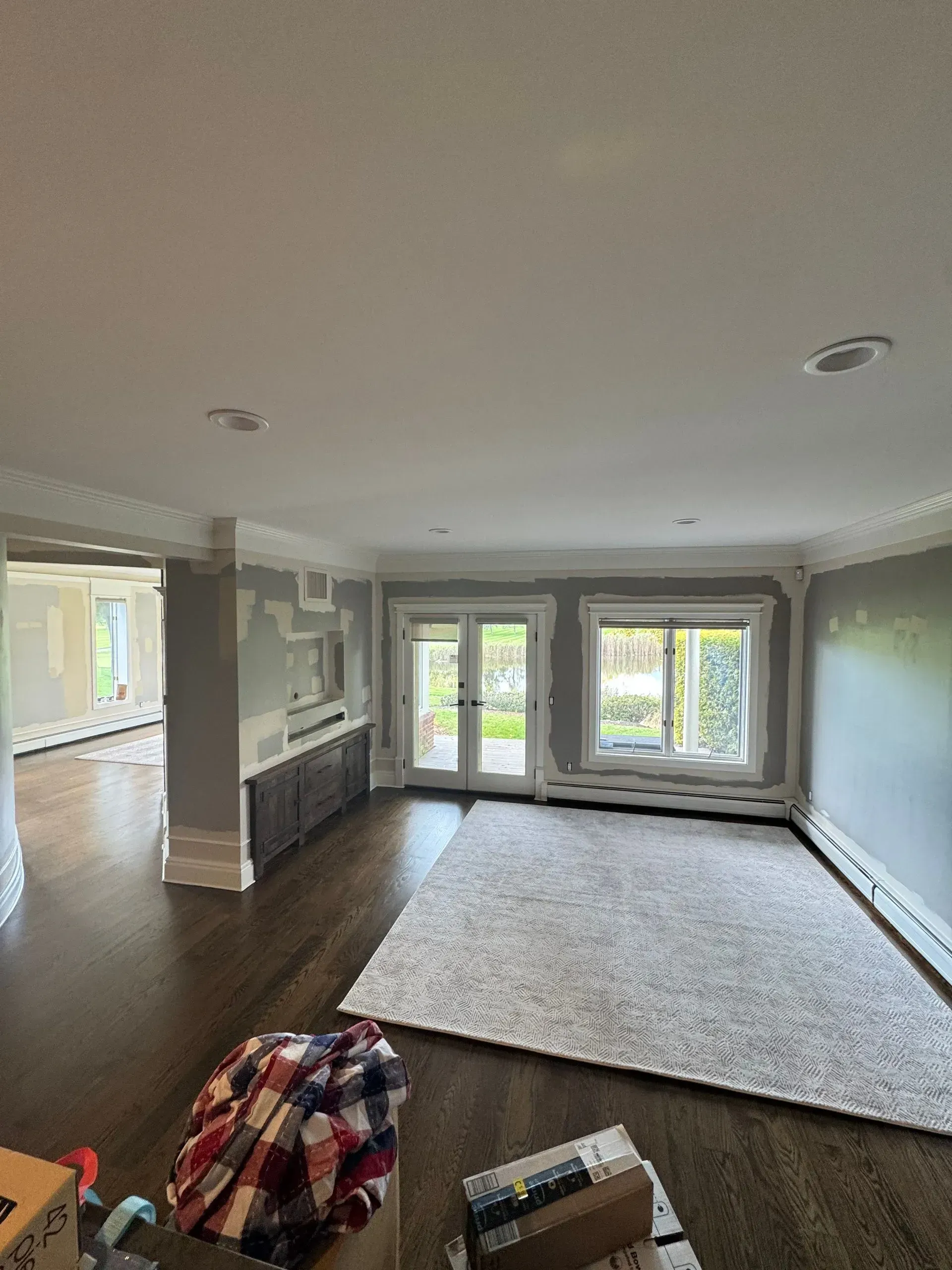 Spacious empty living room with dark wood floors, white rug, and large windows overlooking greenery.