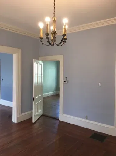 Empty dining room with blue-gray walls, dark wood floor, chandelier, and open doorway to a bright room