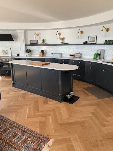 Modern kitchen with dark island, curved cabinetry, herringbone wood floor, and open shelves along the back wall
