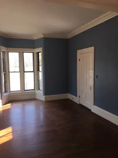 Empty room with dark blue walls, hardwood floor, bay window, and a white door.
