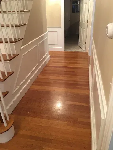 Wooden hallway with stairs on the left, white paneled walls, and an open doorway at the end.