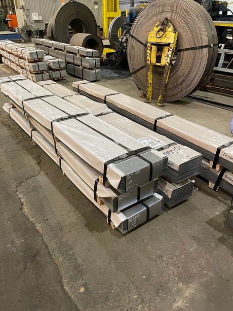 Stacked metal beams strapped on a warehouse floor with large coils and lifting equipment in the background