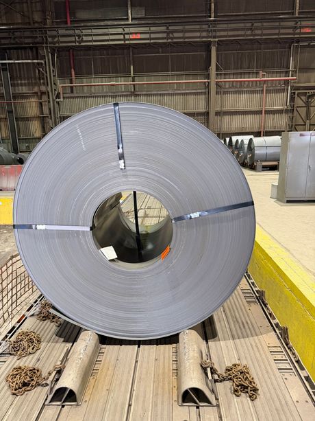 Large silver metal coil on a factory floor with pipes underneath and industrial equipment in the background
