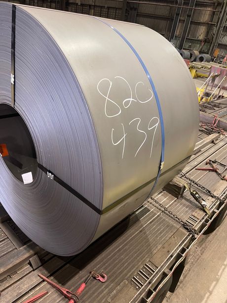 Large rolled steel coil in an industrial warehouse, marked with white chalk and blue straps.