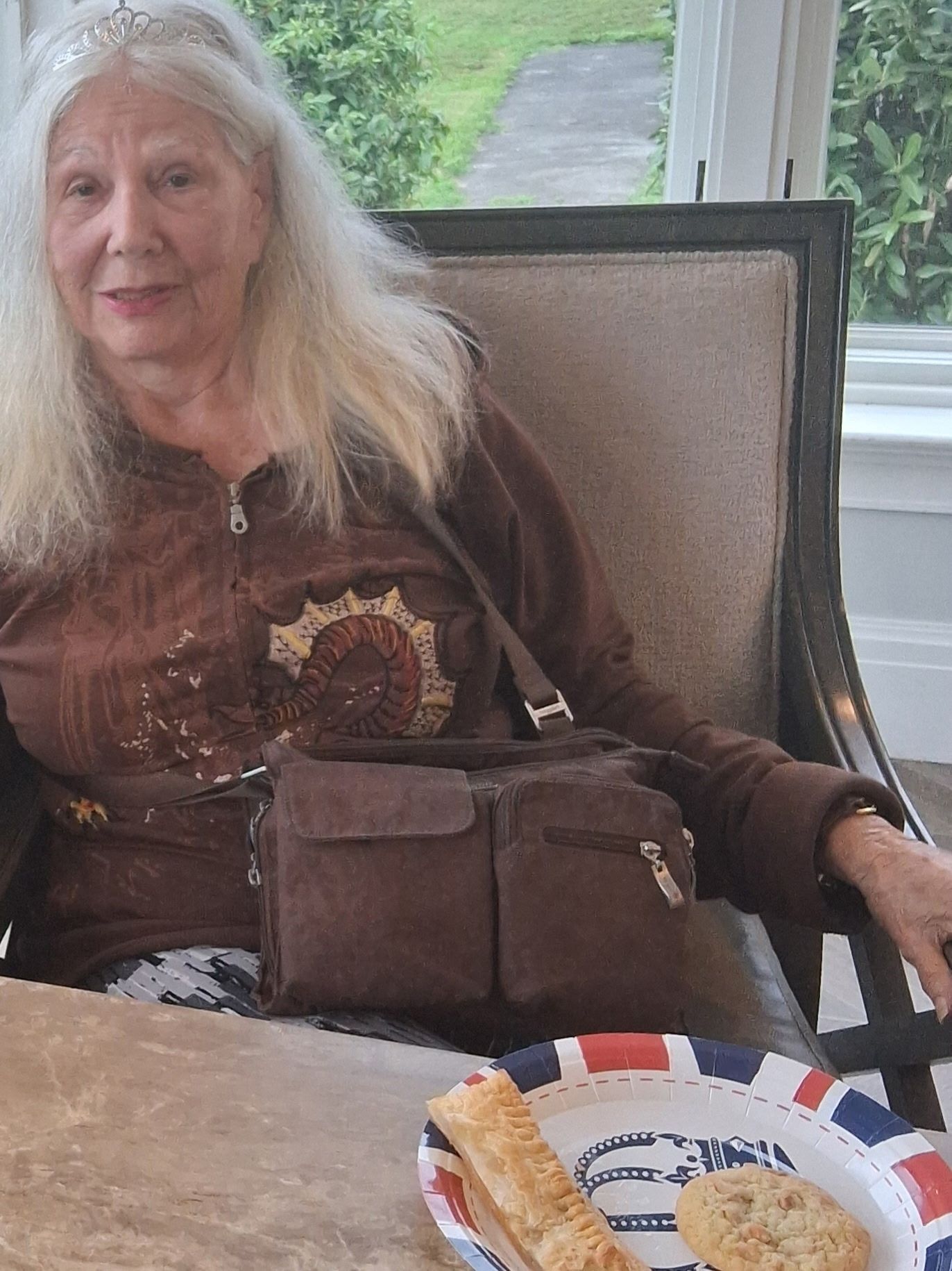 Woman seated indoors at a table with a plate of food, wearing a brown shirt and looking toward the camera
