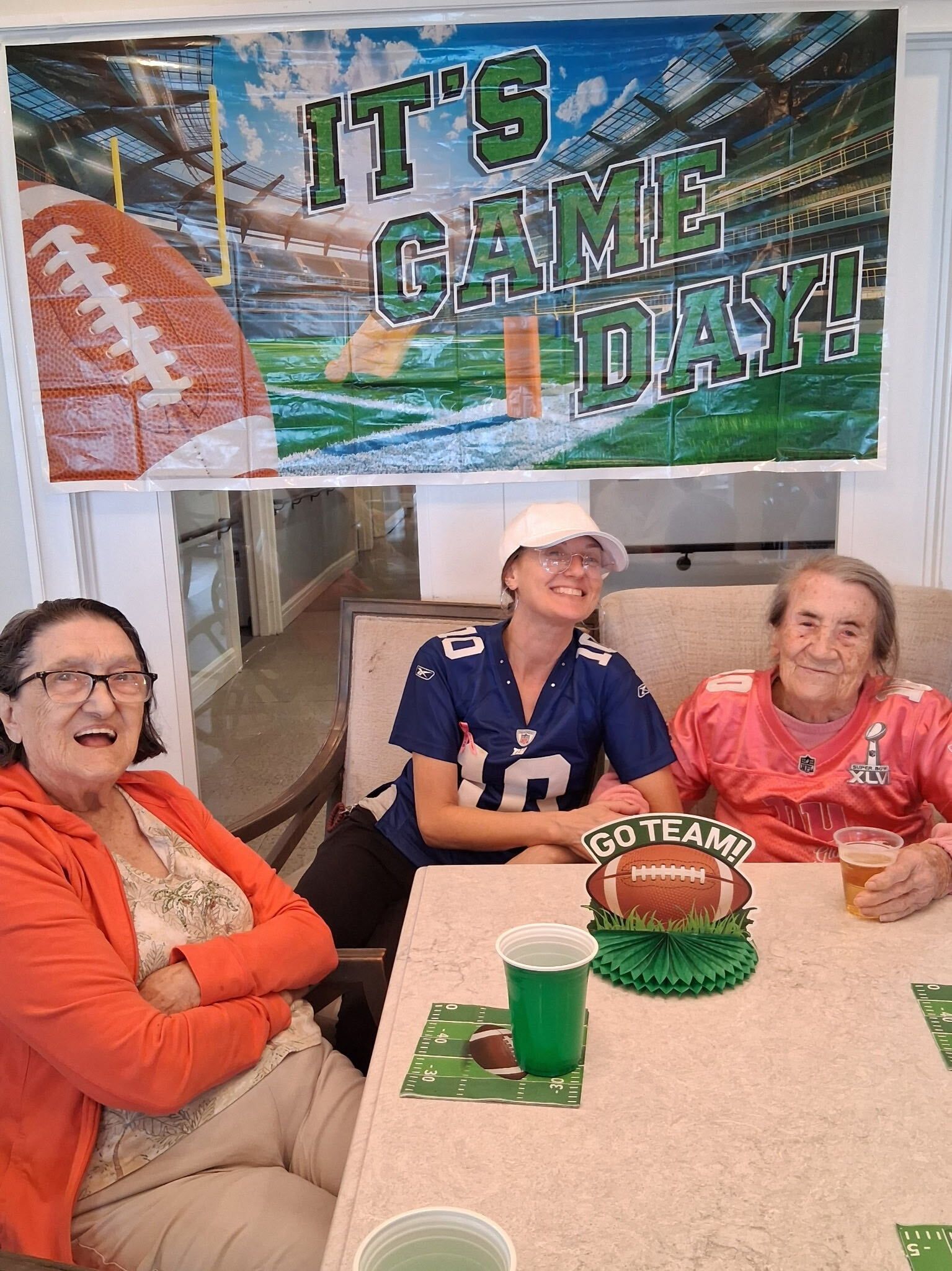 Three women sitting at a table under an “It’s Game Day!” banner, smiling with green drinks and football decor.