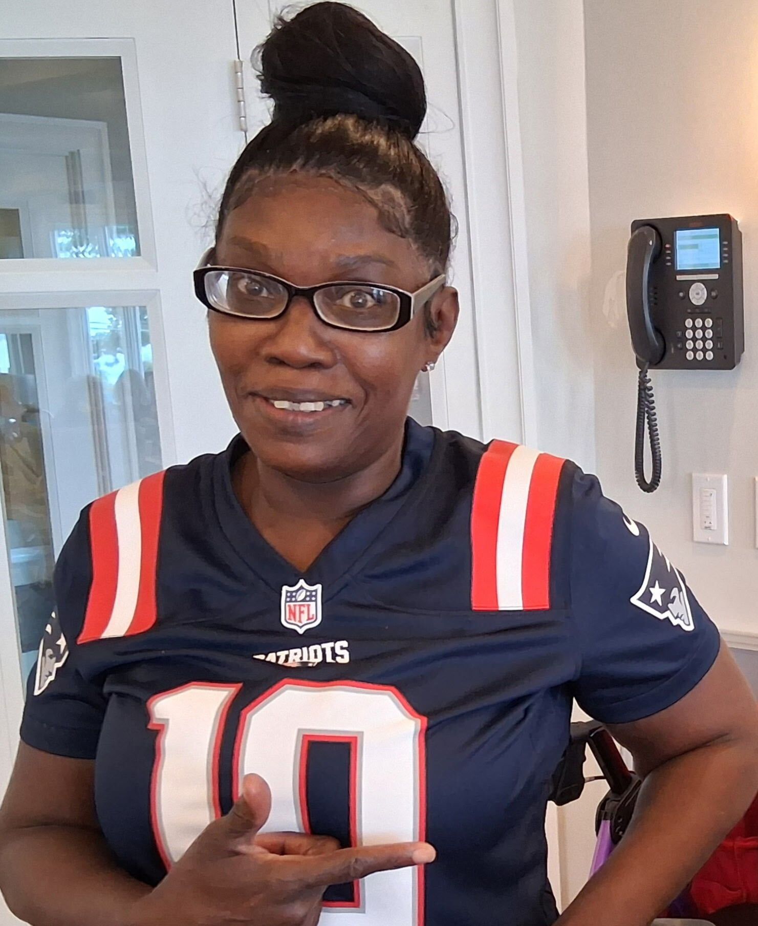 Woman in a navy sports jersey with a high bun and glasses, smiling indoors and making a hand gesture.