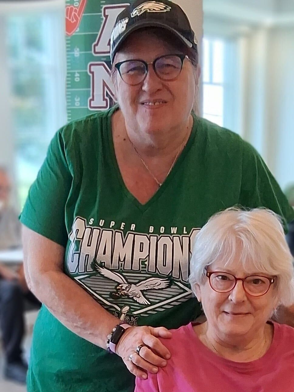 Two women posing together indoors, one standing behind the other, both smiling and wearing glasses.