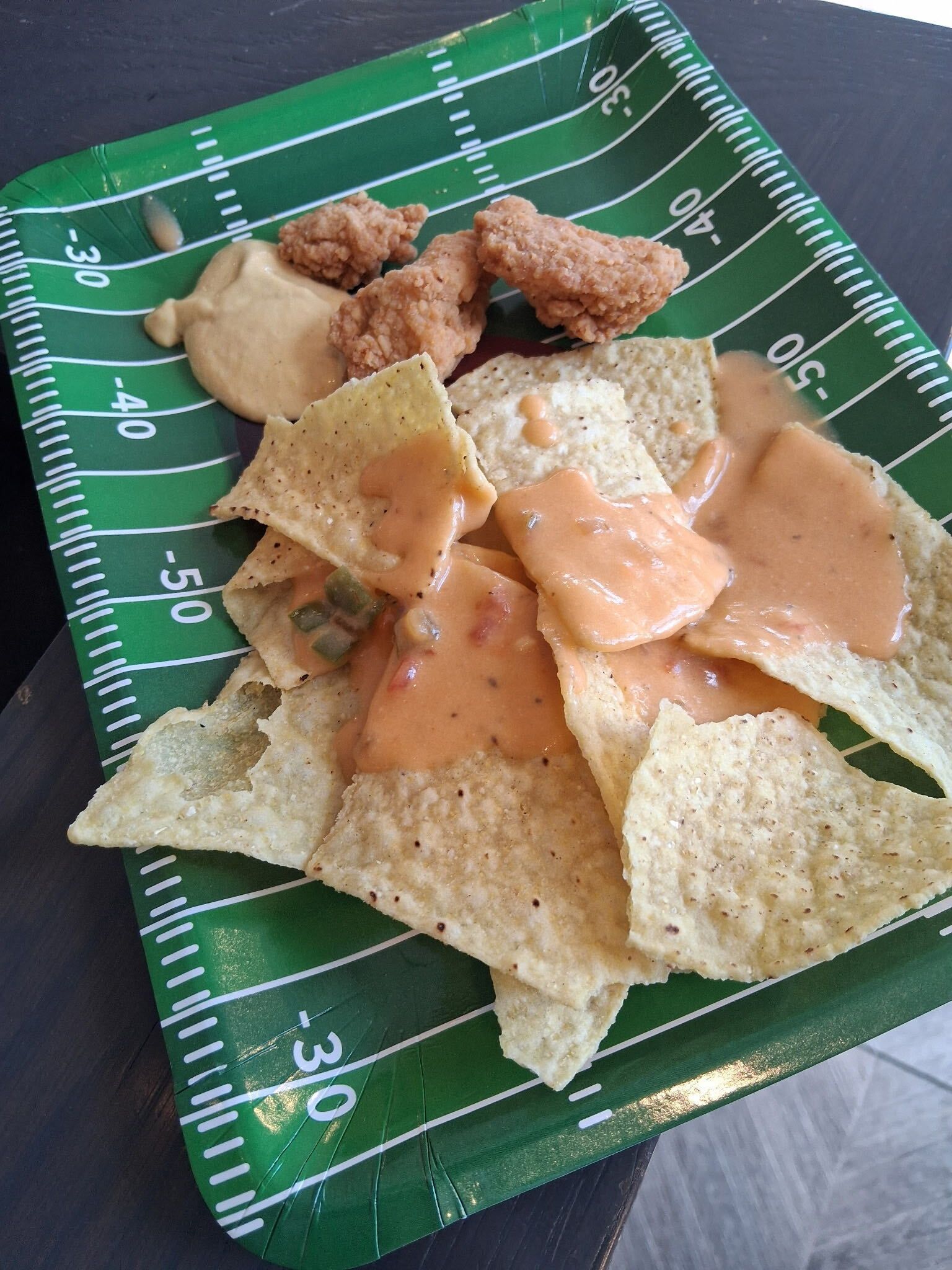 Tortilla chips and small chicken pieces on a green football field serving tray with dipping sauce
