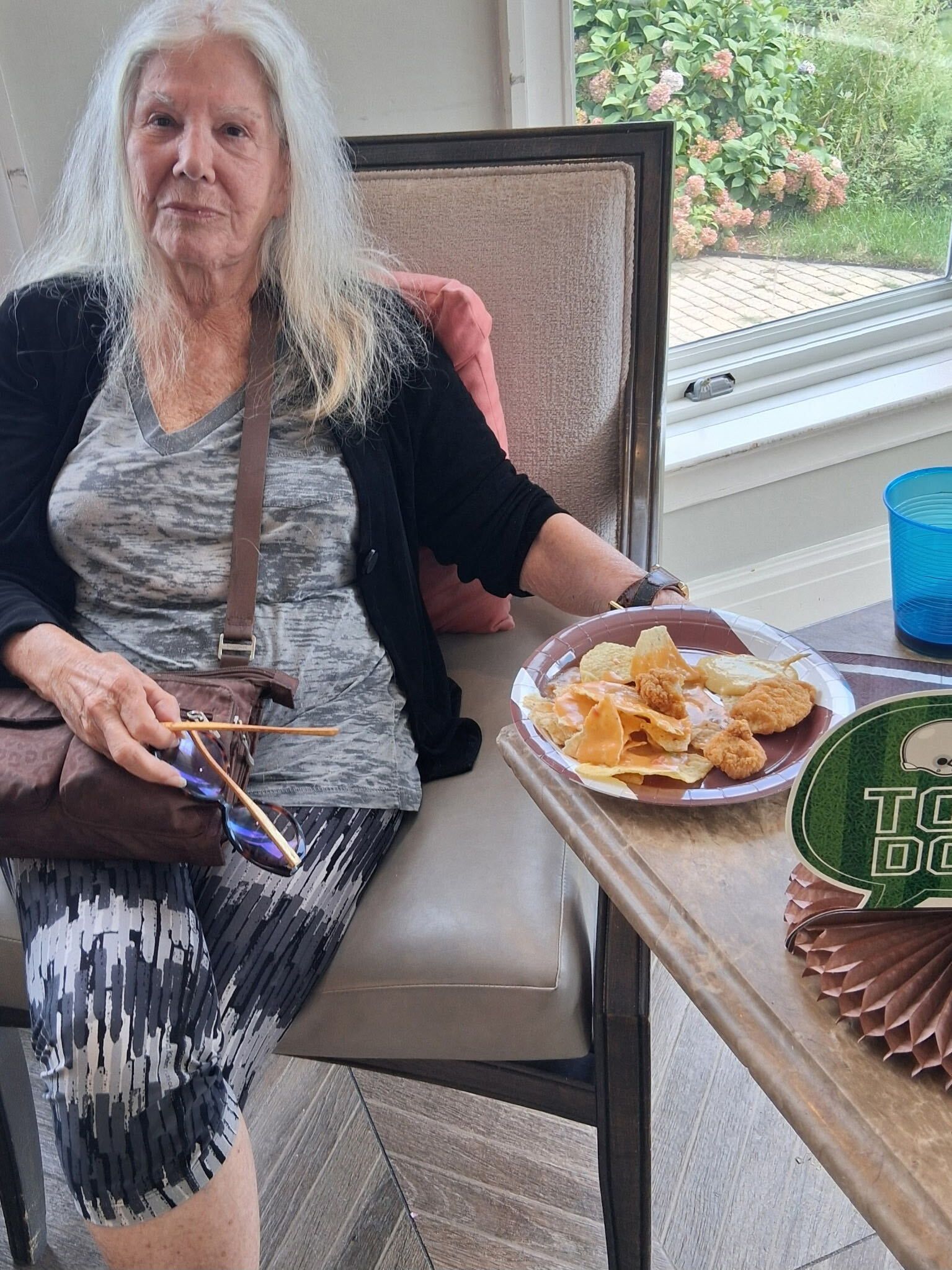 Woman seated on a patio chair holding a drink, beside a table with snacks and a blue cup.
