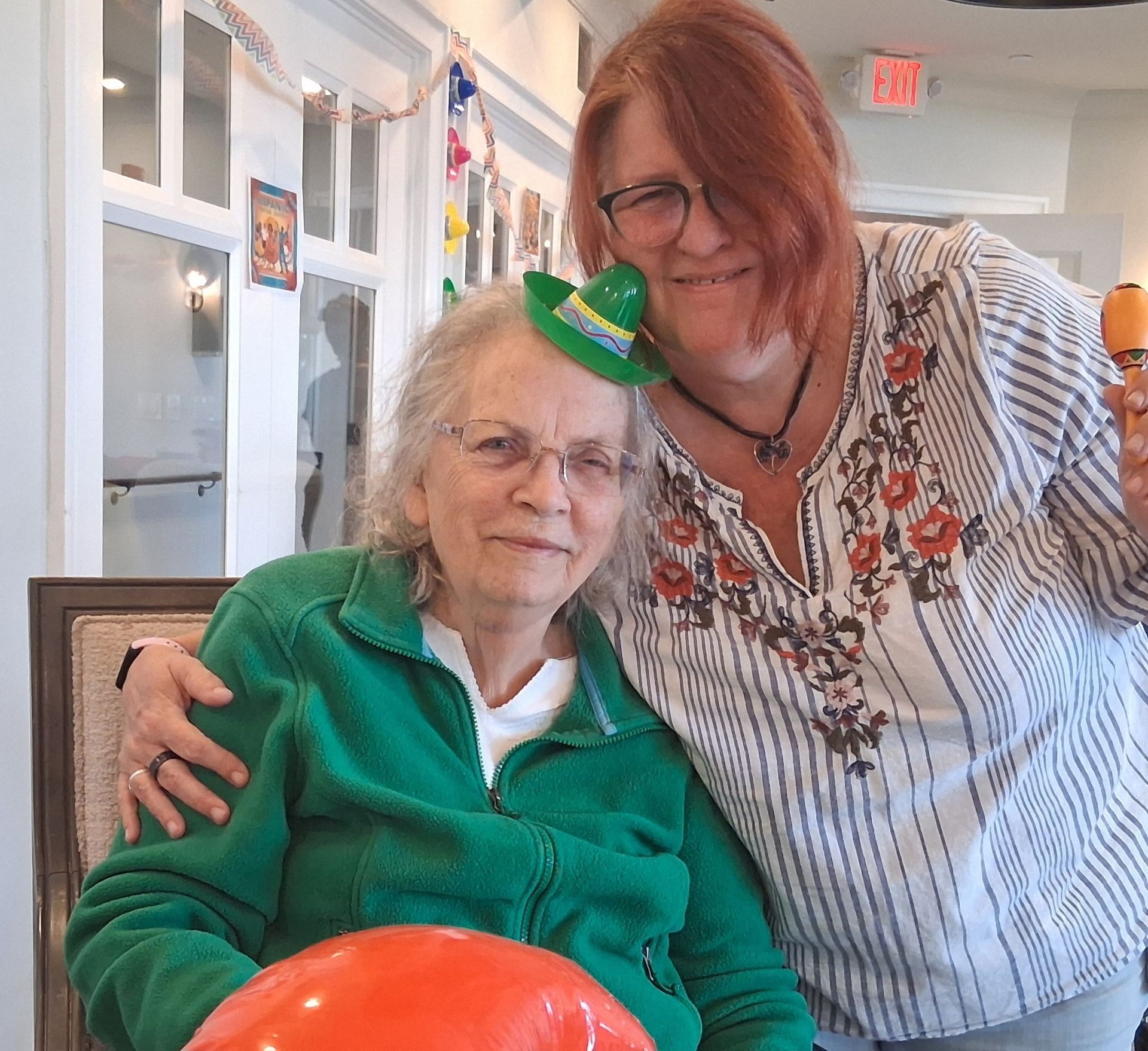 Two women smiling indoors, one seated in green, the other standing beside her with an arm around her shoulders.