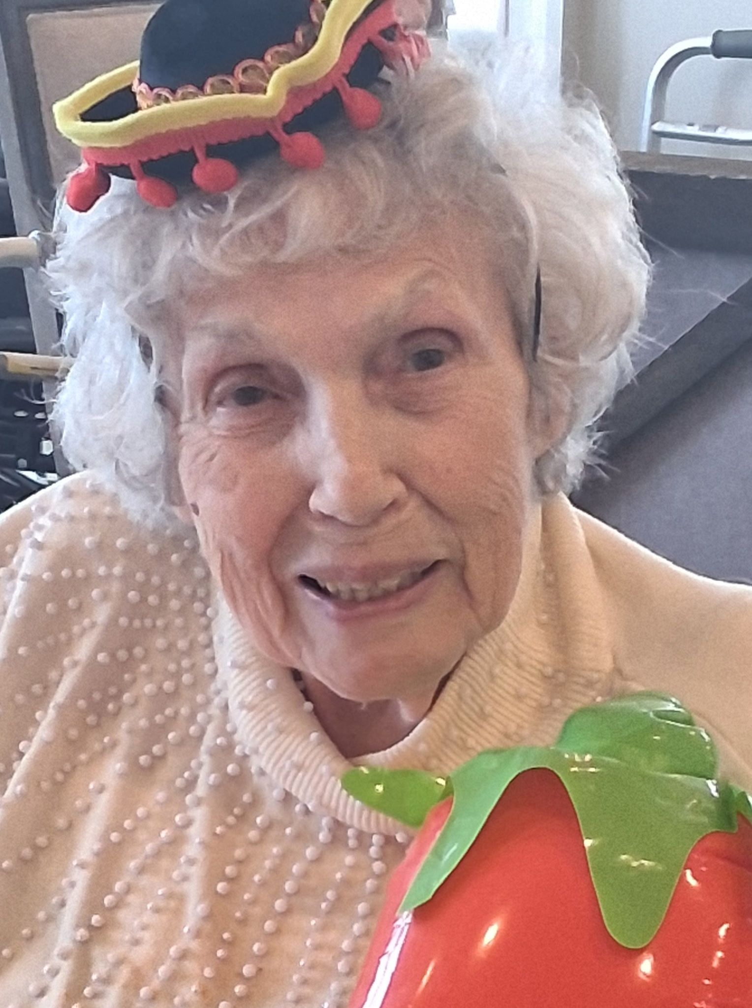 Smiling person wearing a small hat and holding a colorful strawberry-shaped prop indoors