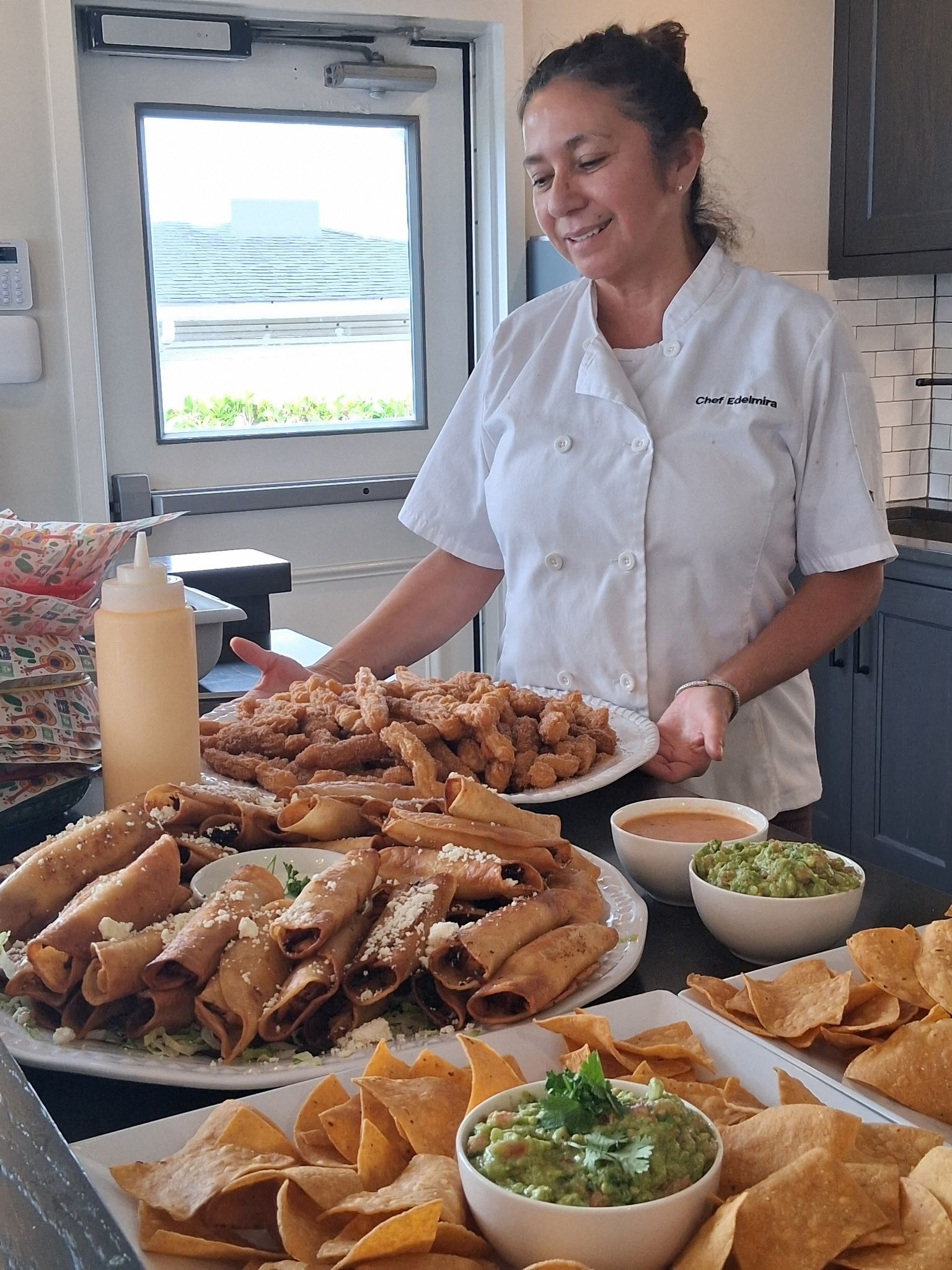 Smiling chef serving a buffet of fried snacks, dips, and chips in a bright kitchen