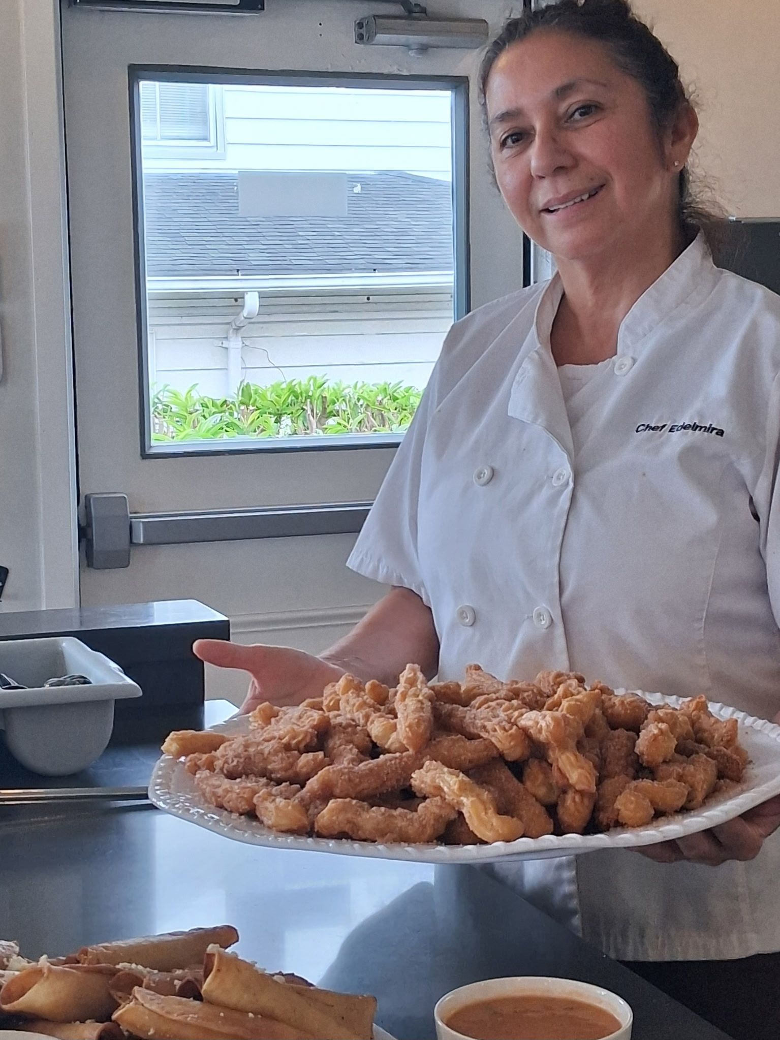 Person in a white chef coat holding a tray of fried food in a kitchen.