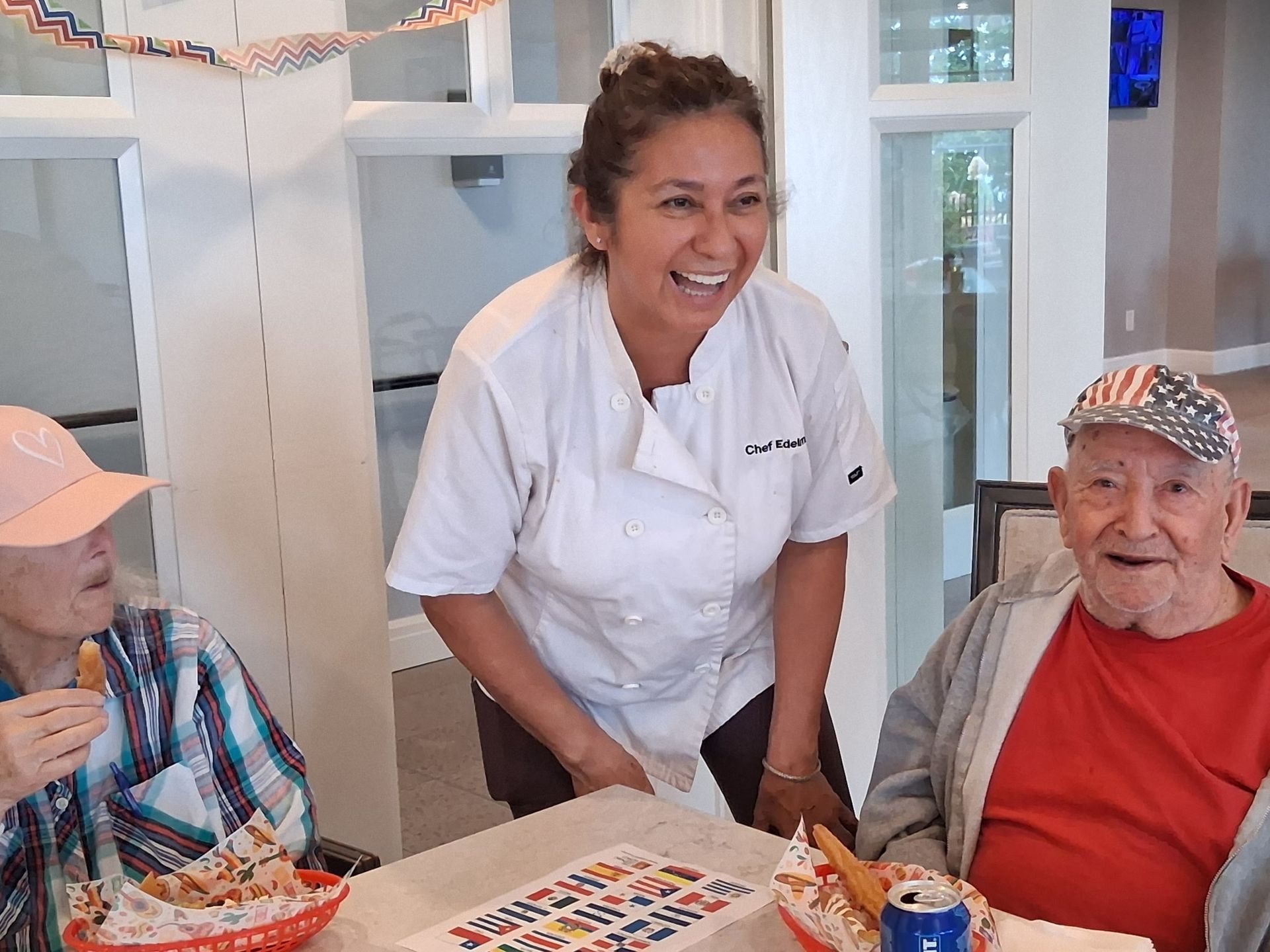 Smiling chef leaning toward two seated diners at a table in a bright dining room