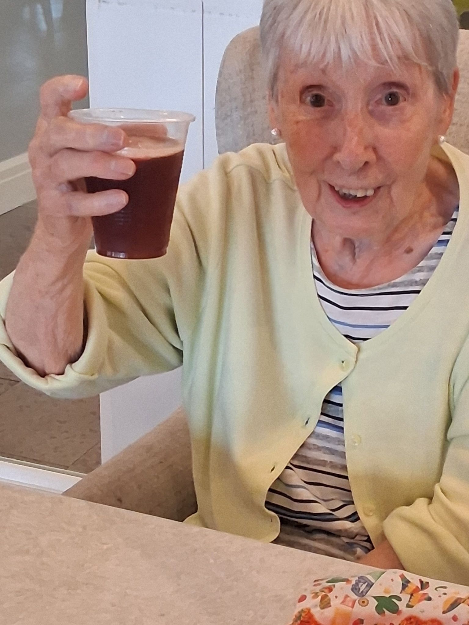 Smiling person holding up a cup of iced drink at a table, wearing a pale yellow cardigan.