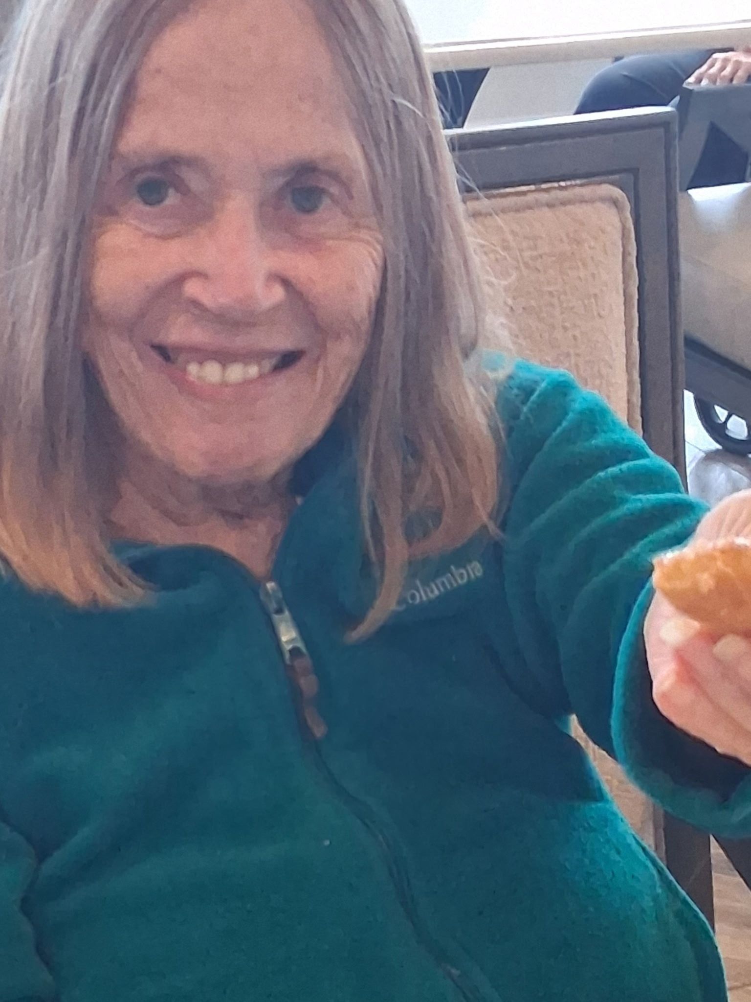 Smiling woman in a teal fleece holds out a pastry while seated indoors