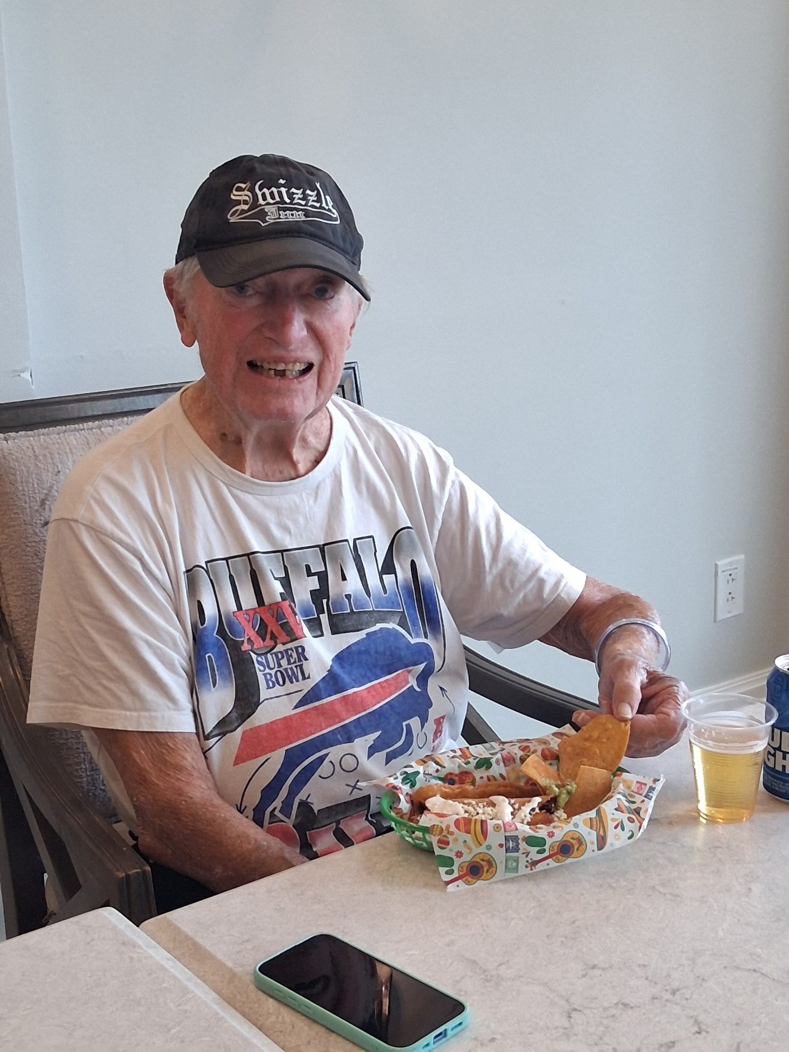 Man eating lunch at a table, wearing a cap and white T-shirt, with a drink and smartphone nearby.