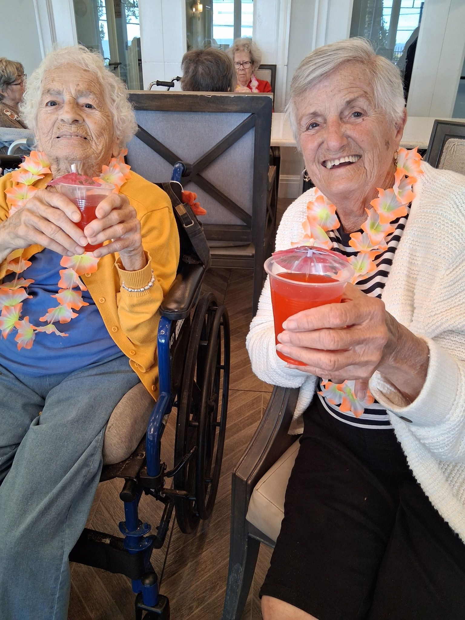 Two smiling residents in wheelchairs holding colorful drinks in a bright common room