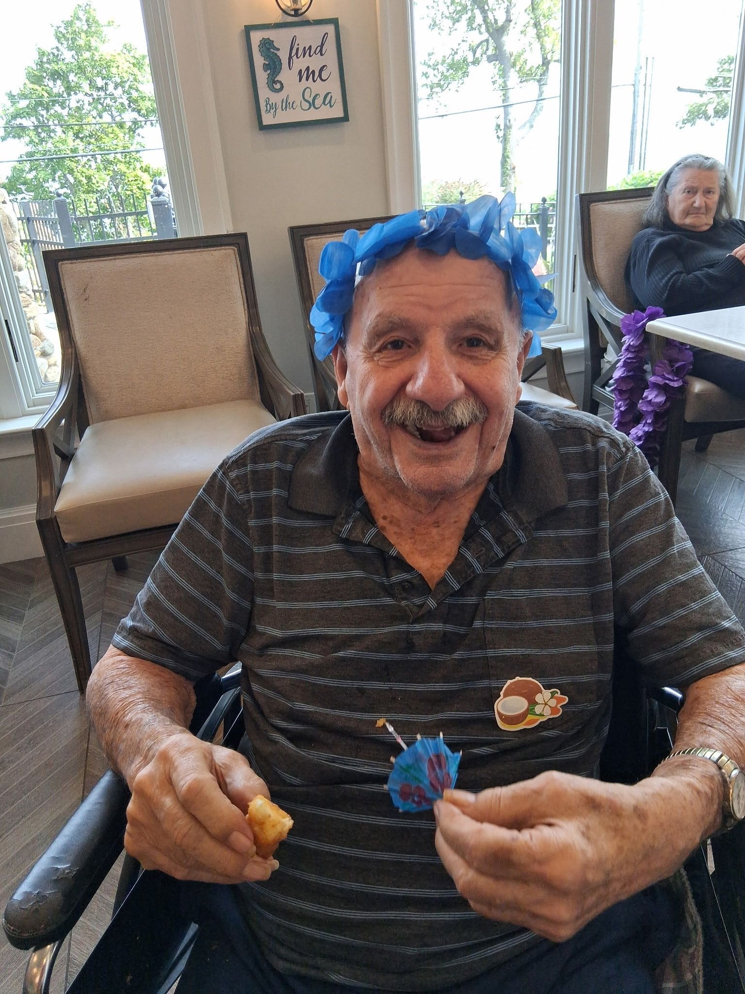 Smiling man in striped shirt with blue flower headband, holding flowers at a table in a bright room