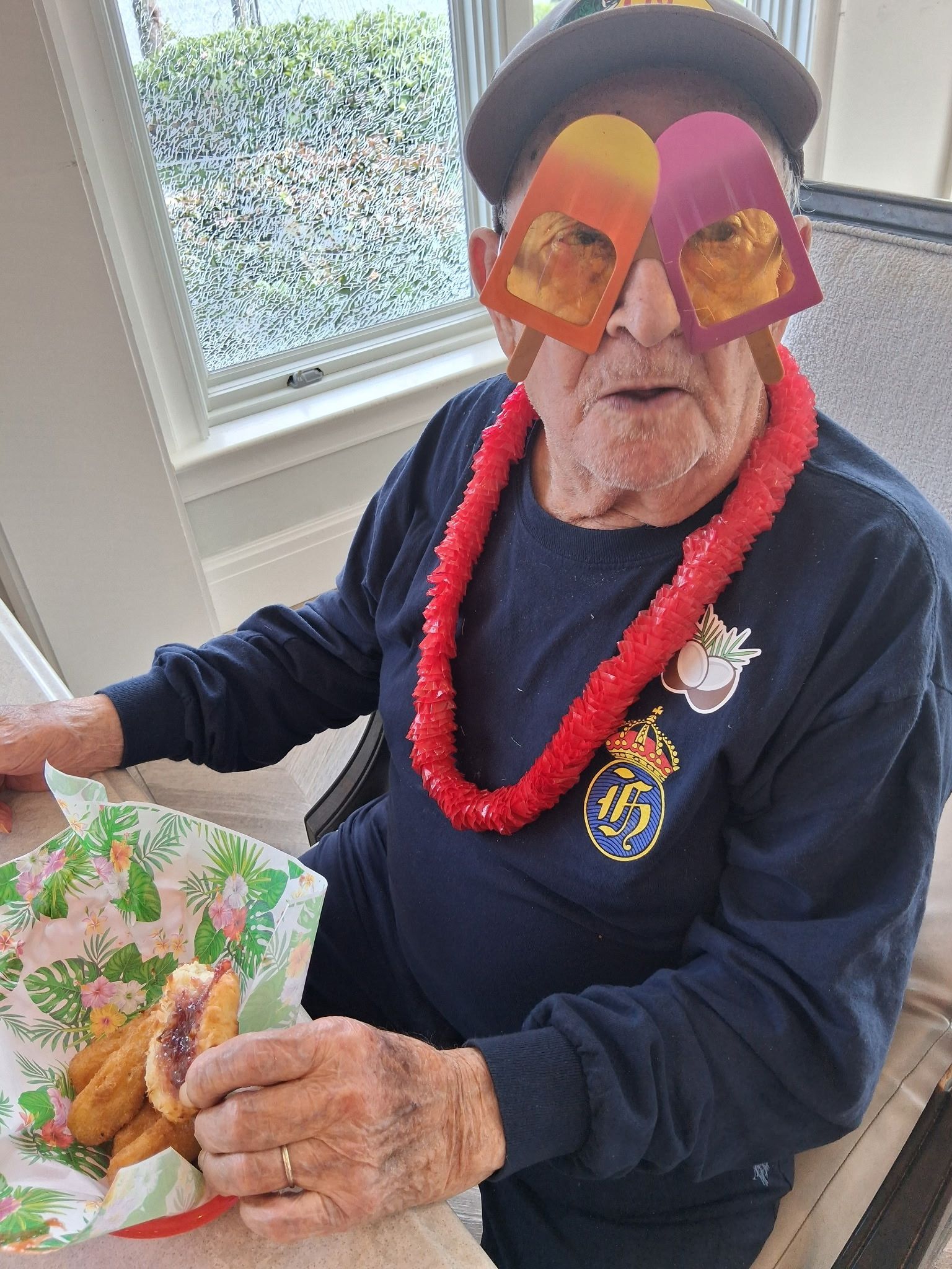 Person wearing a colorful mask and lei, holding a floral card near a window.