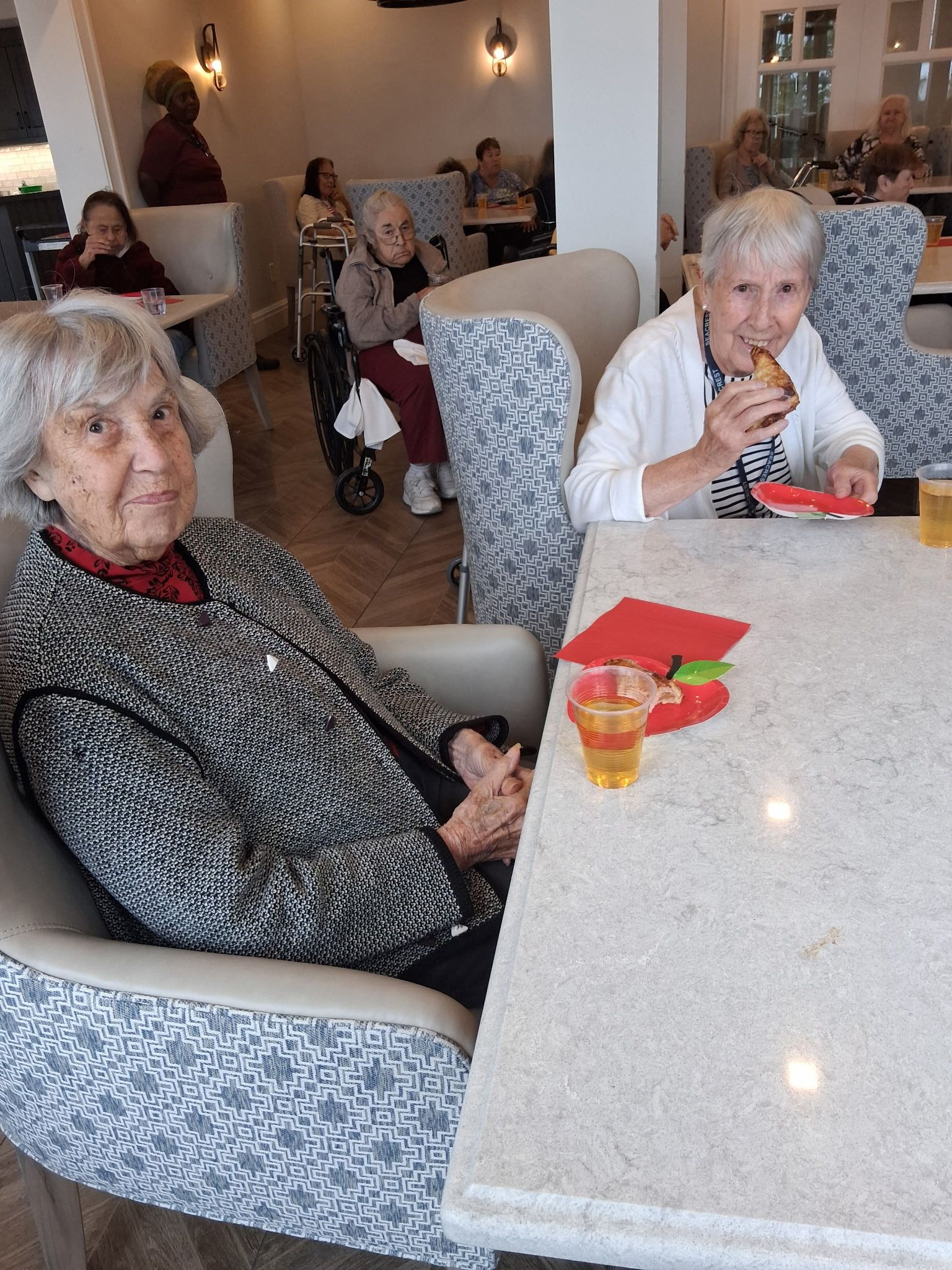 Elderly people seated at a dining table in a bright room, one eating a snack while others chat nearby.