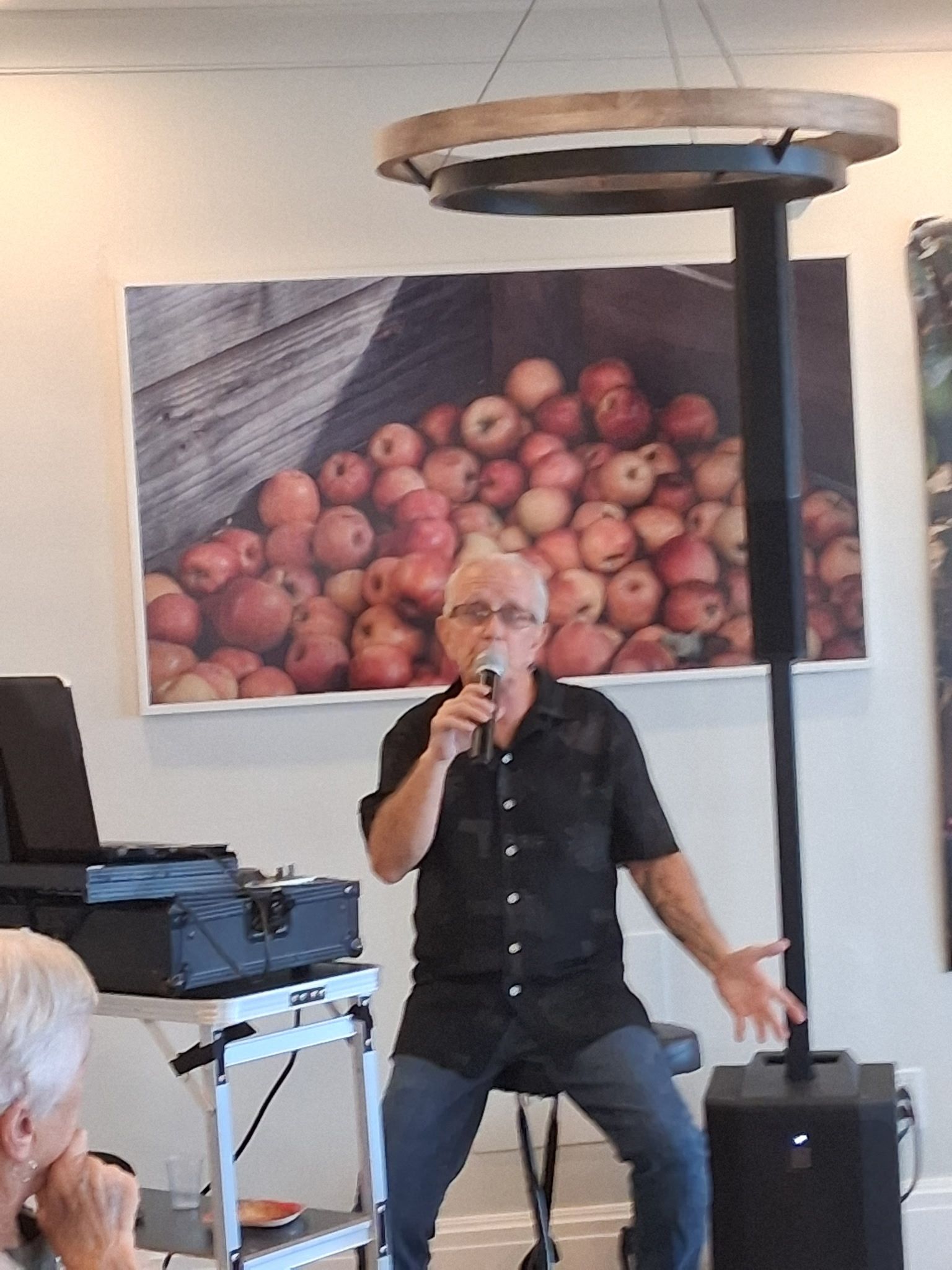 Man speaking into a microphone on a stool before a fruit mural in a casual indoor setting