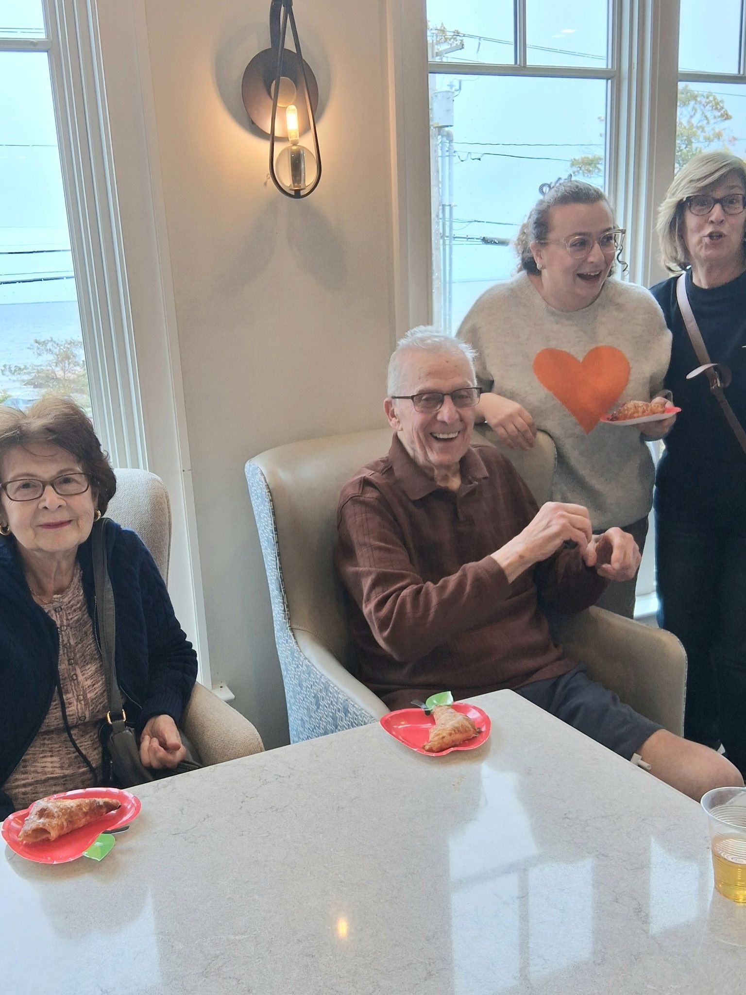 Four people seated at a table in a bright room, smiling and chatting with plates of snacks.