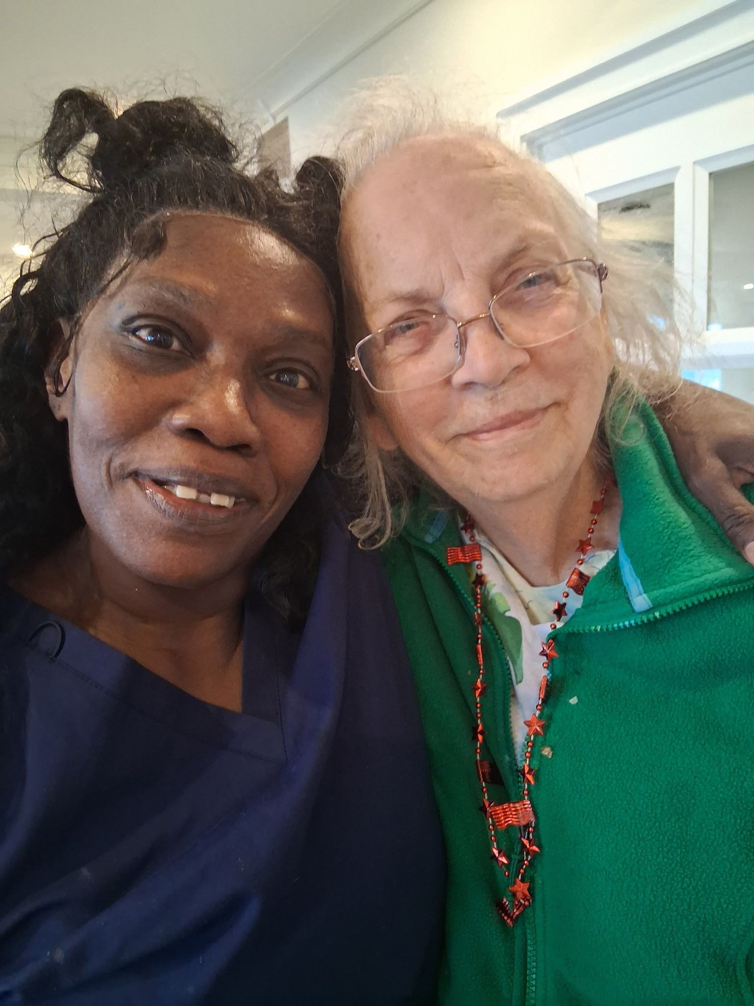 Two smiling people posing closely indoors, one in blue and one in green, near a window.