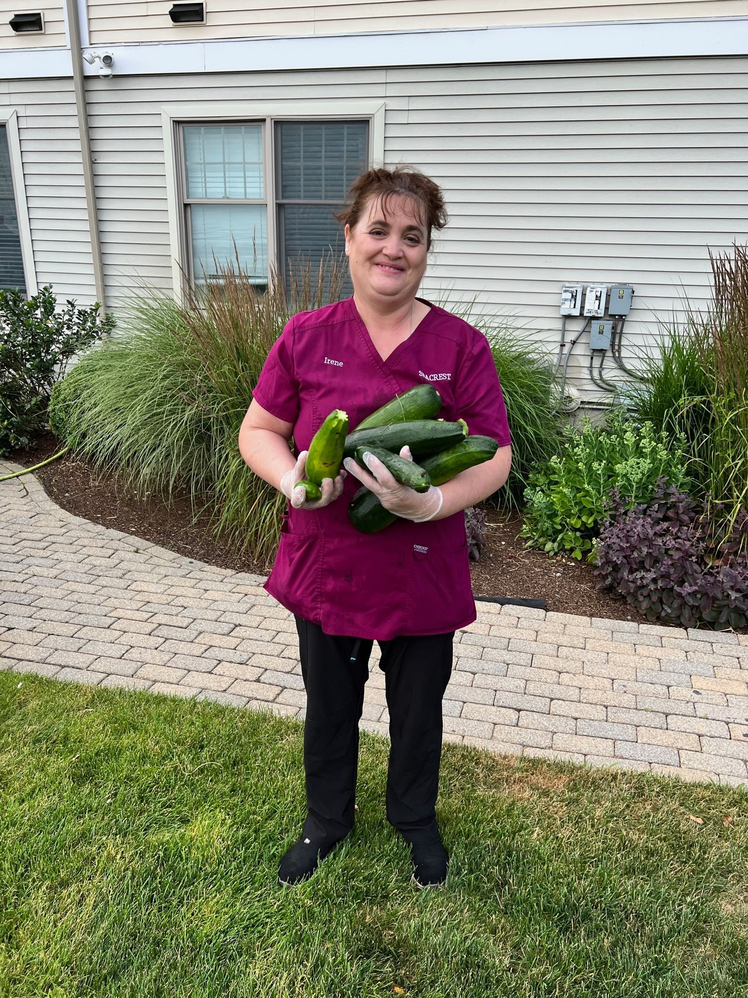 Woman in maroon scrubs holds several large cucumbers outside a building with bushes.