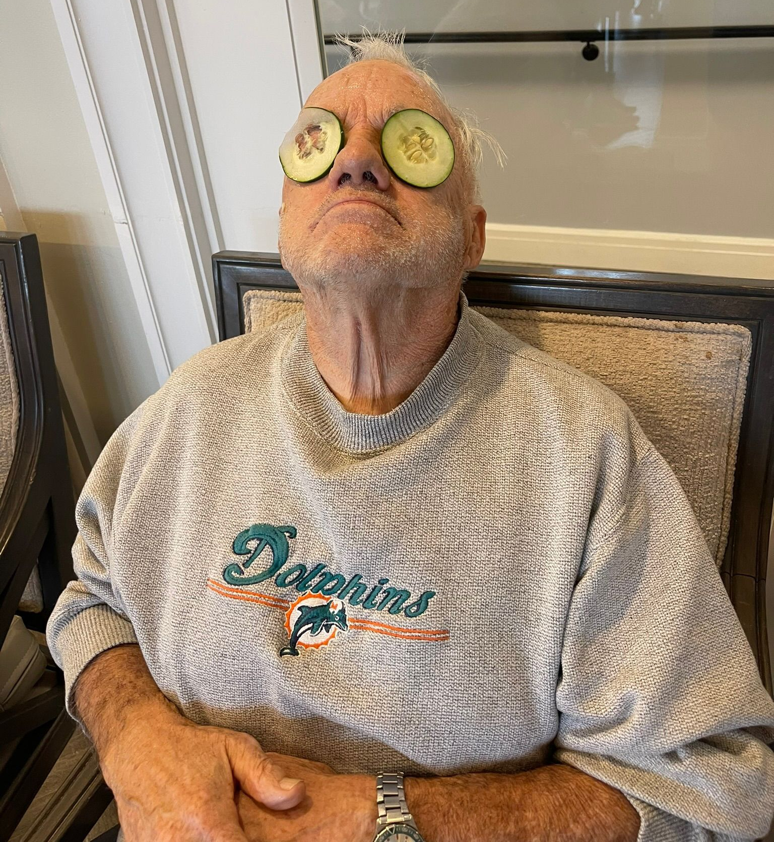 Man with cucumber slices on eyes, wearing a Dolphins sweatshirt, sitting in a chair.