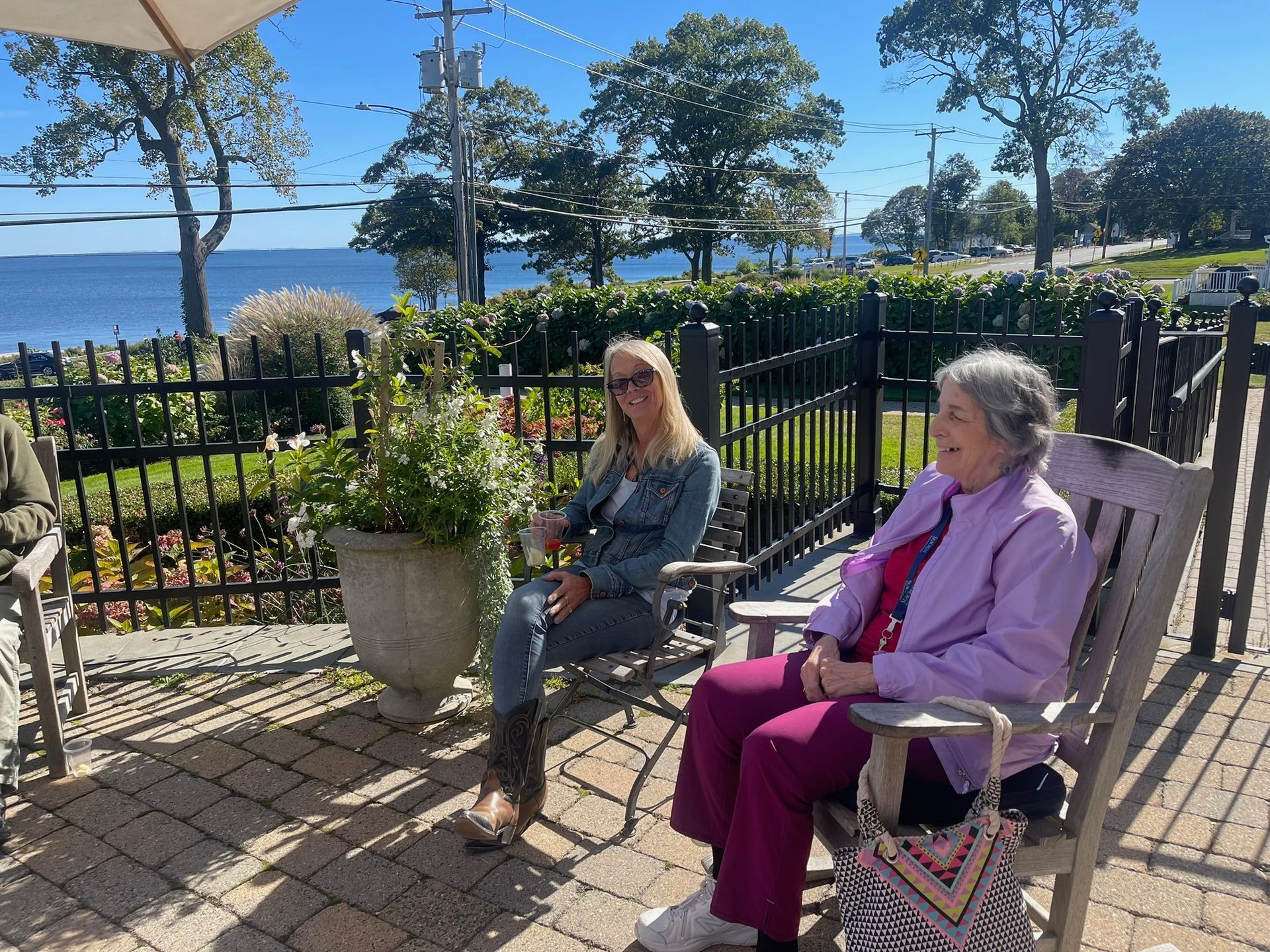 Two people seated on a patio, overlooking a body of water. One wears a denim jacket, the other a purple jacket.