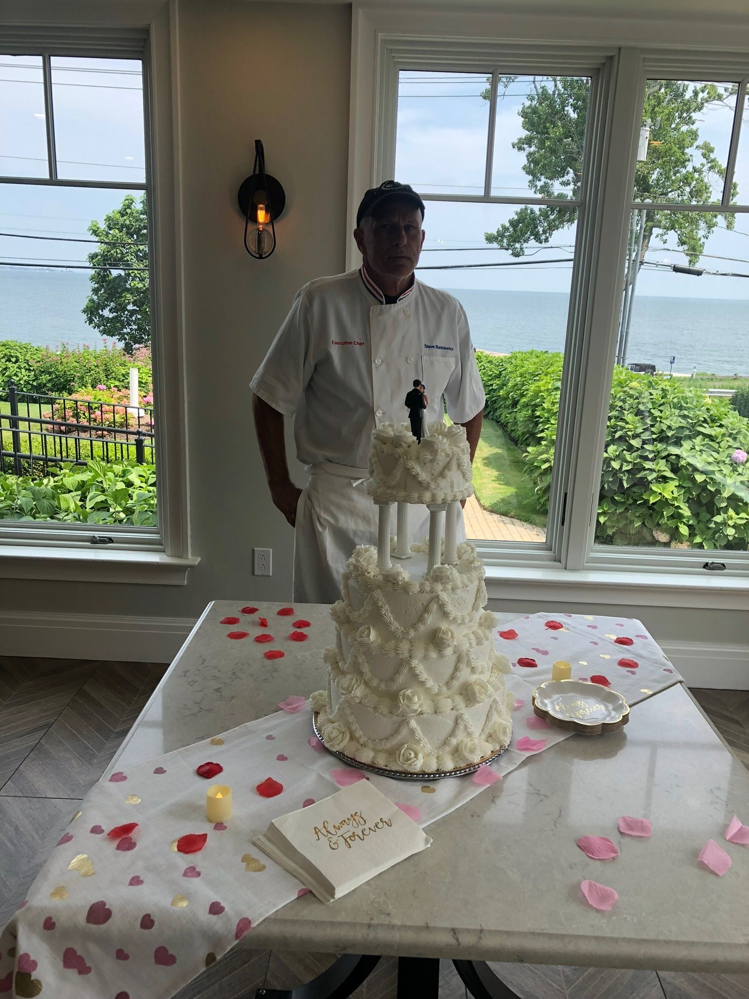 Chef standing by a three-tiered wedding cake on a table. The cake has white frosting and a couple figurine on top.