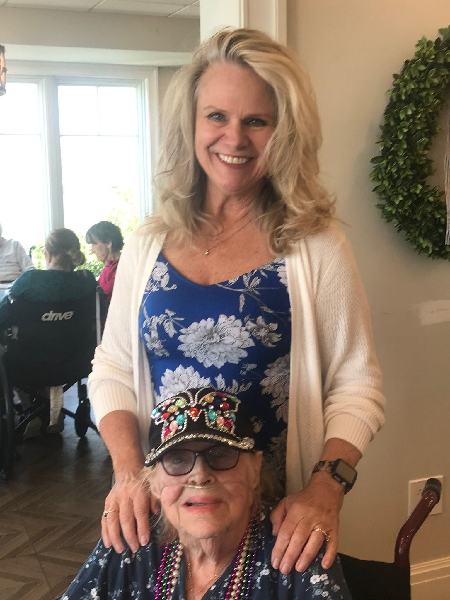 Woman smiling behind an elderly woman in a hat. Both are indoors, with people and decorations in the background.