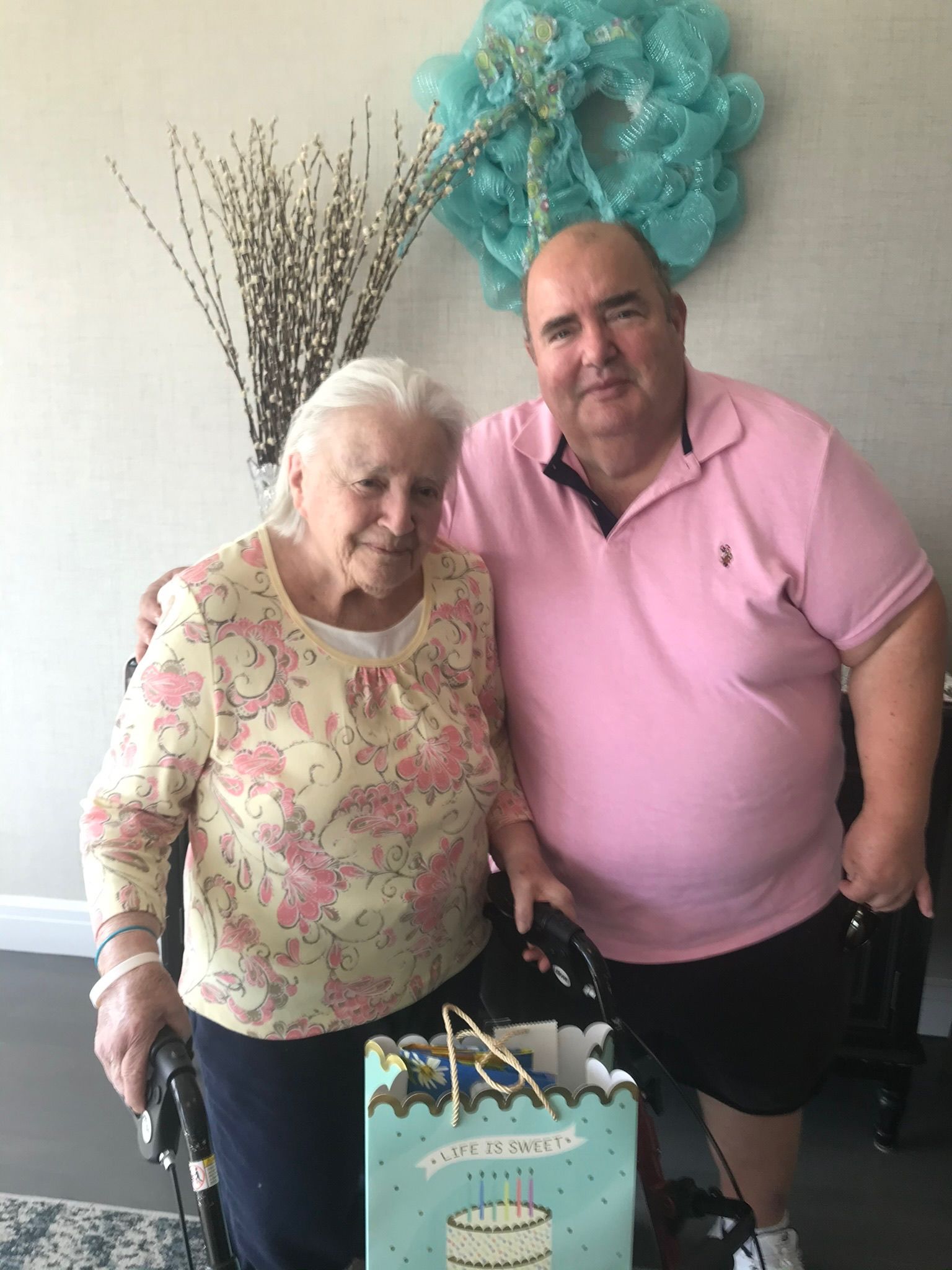 Elderly woman with walker and man with arm around her, holding a gift bag. Light pink shirt, floral top, and decorative wreath.