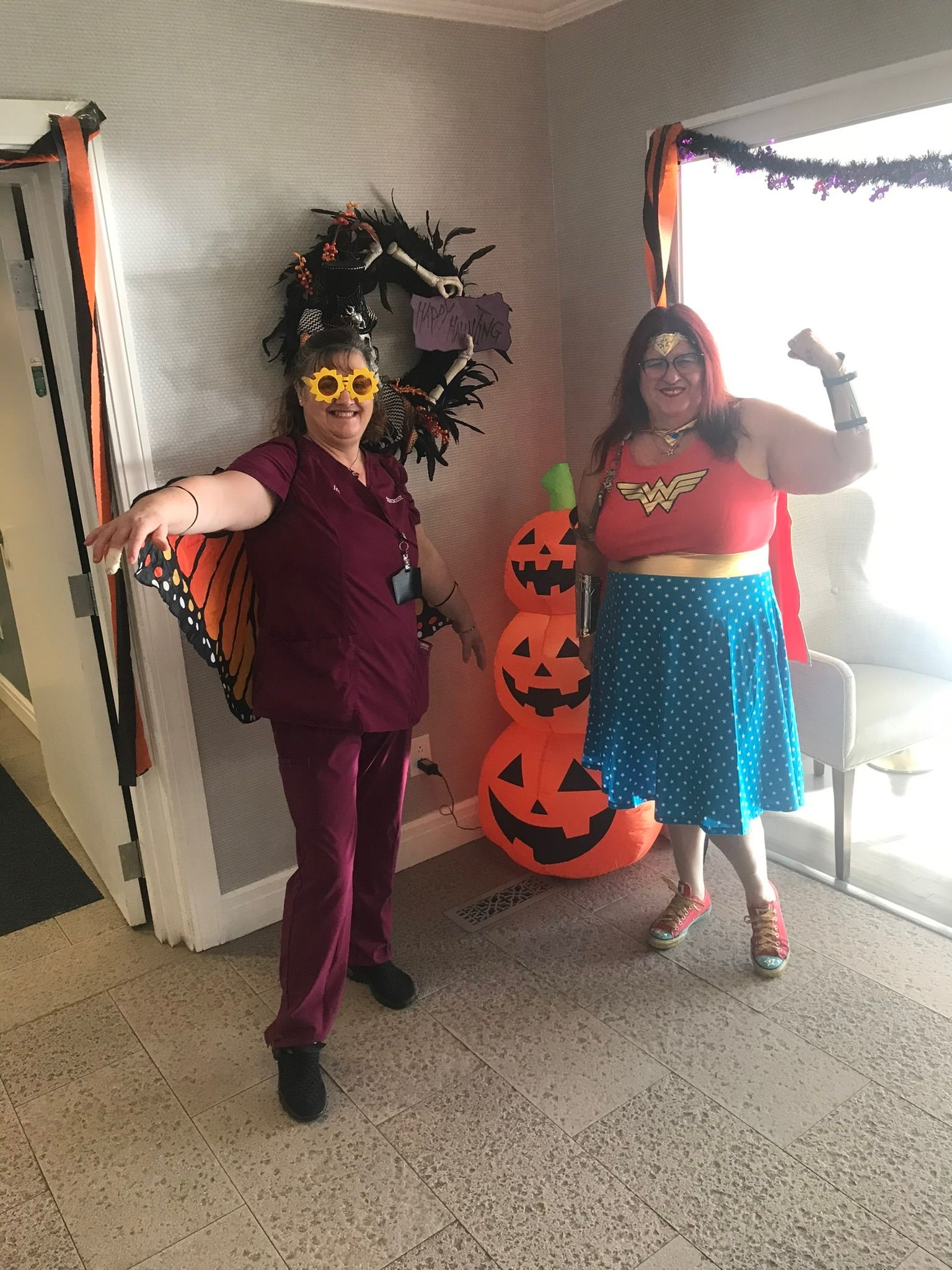 Two women in Halloween costumes pose in front of decorations; one wears butterfly wings and the other Wonder Woman attire.
