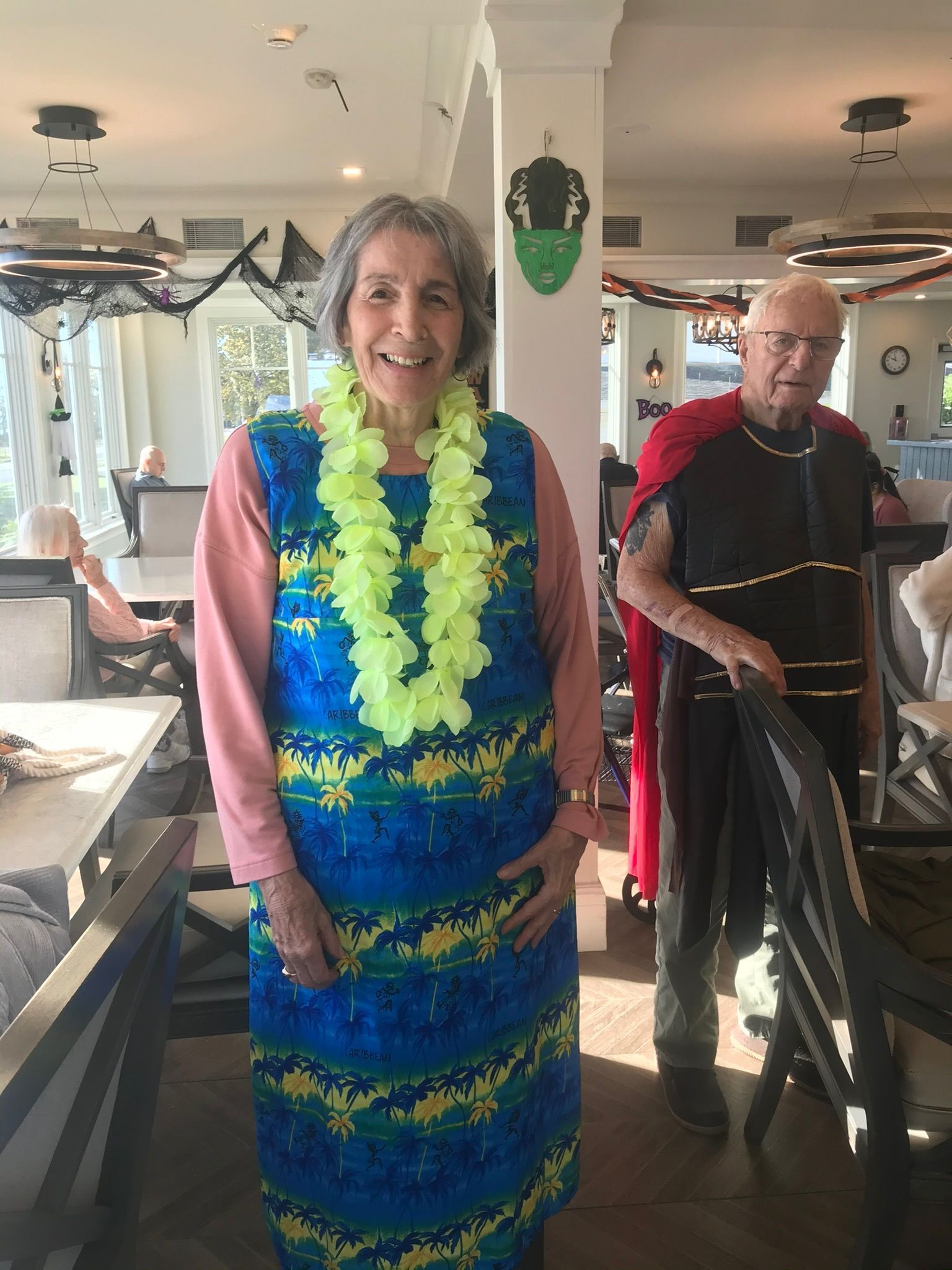 Woman in blue dress and lei smiles; man in Roman attire holds chair, both inside a decorated room.