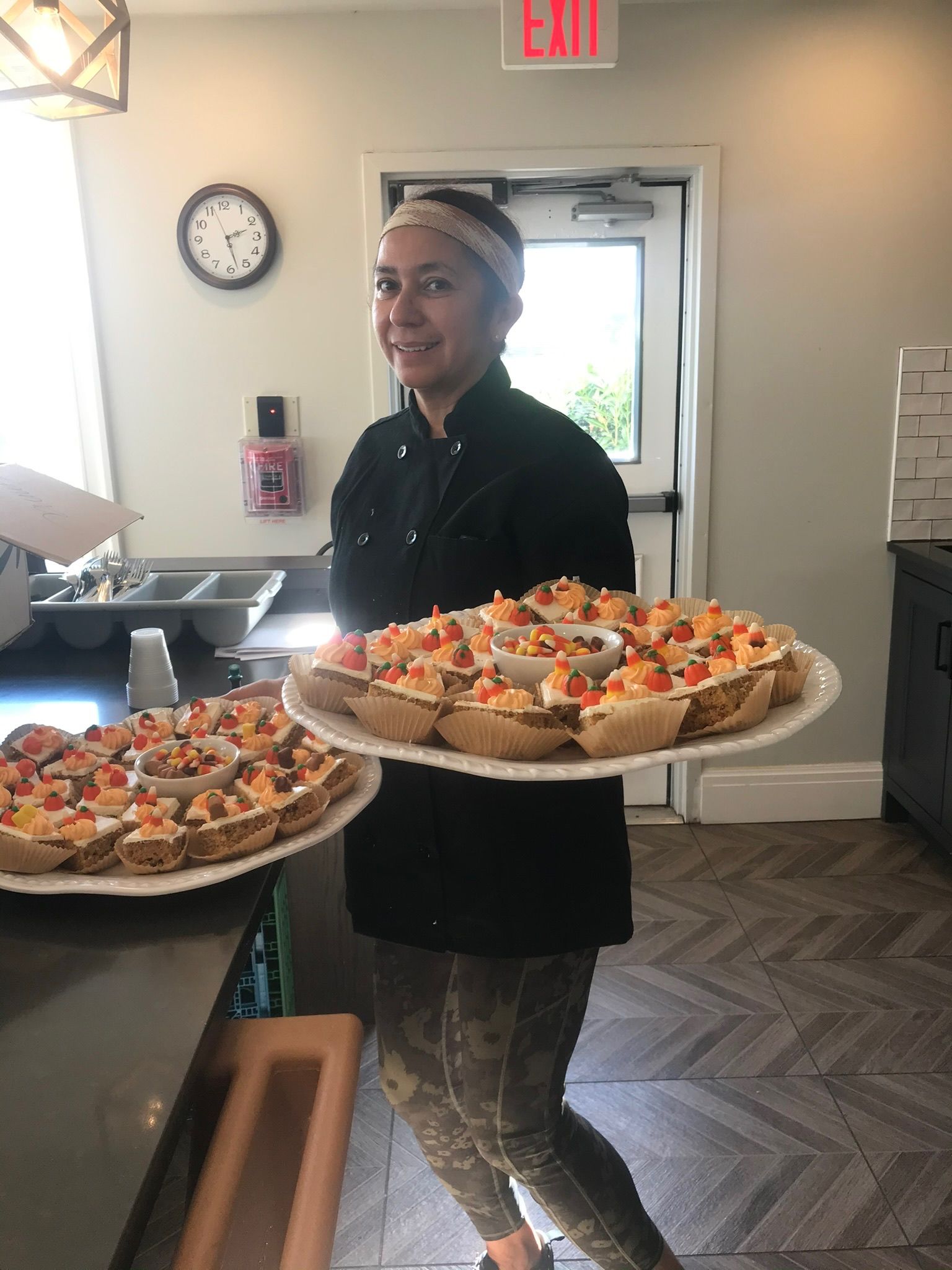 Woman in black chef's coat holds two platters of appetizers in a kitchen. She smiles, with a clock and exit sign visible.