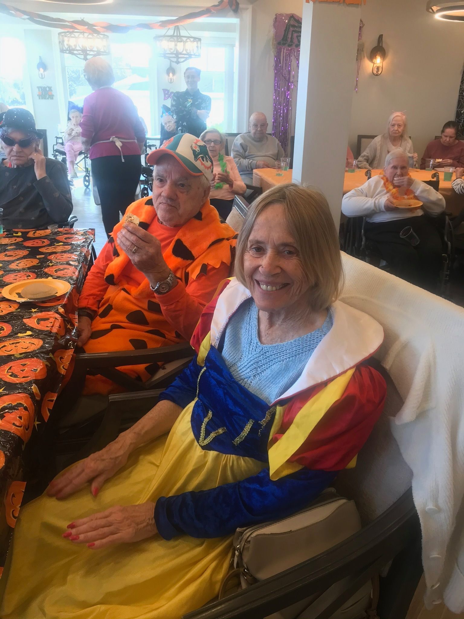 People in Halloween costumes at a gathering. Woman in Snow White dress smiles. Man in pumpkin costume eats.