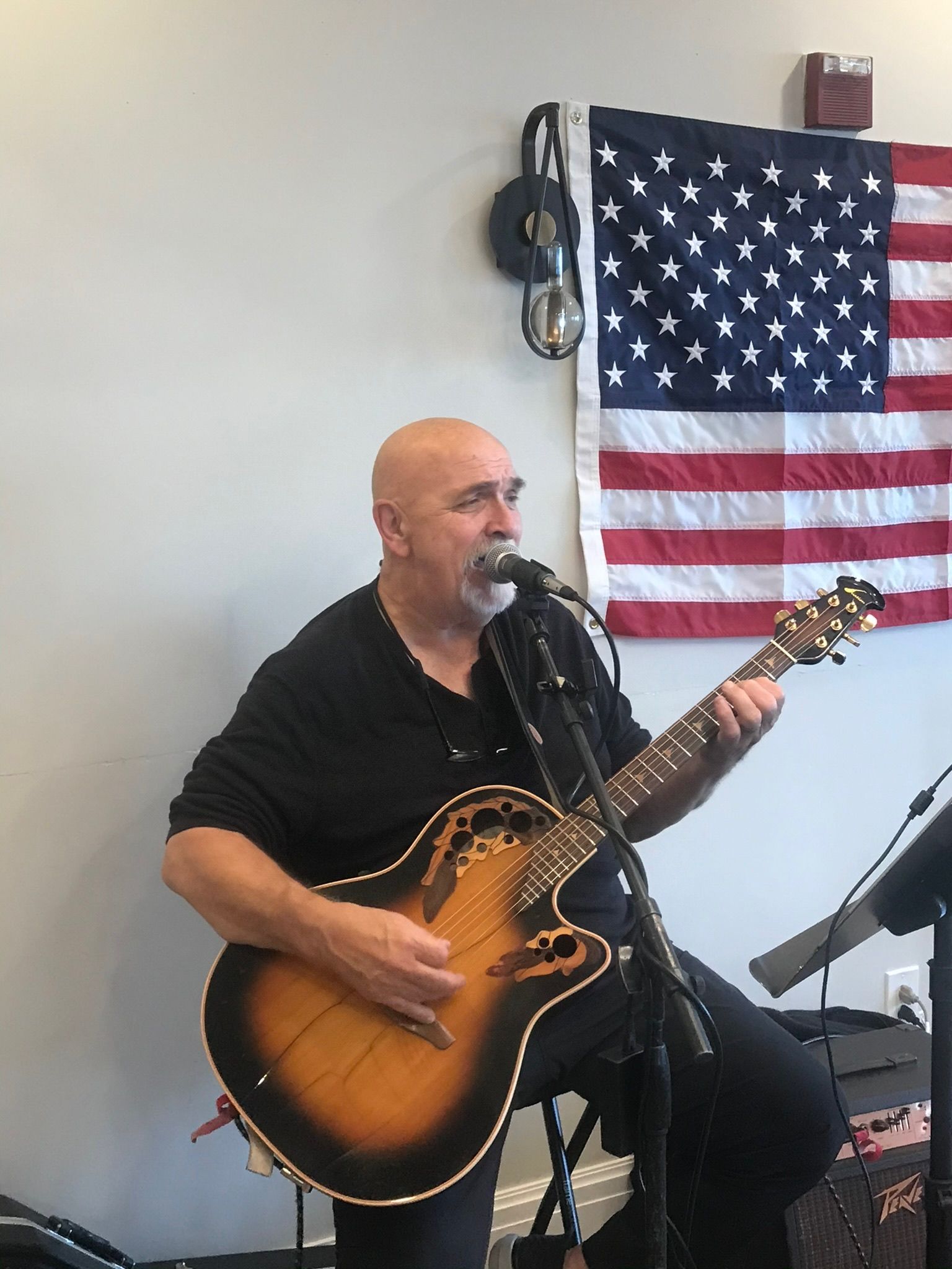 Bald man plays guitar, singing into a microphone, American flag in the background.