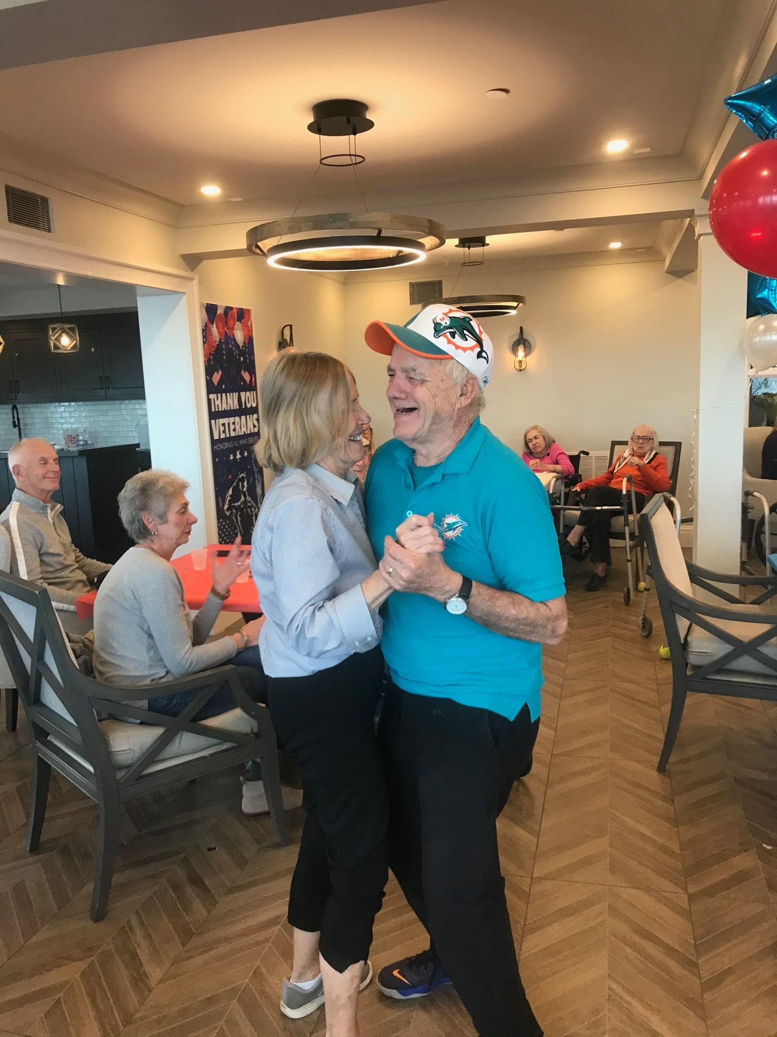 Elderly couple dancing in a brightly lit room; they smile and hold hands. Others watch nearby.