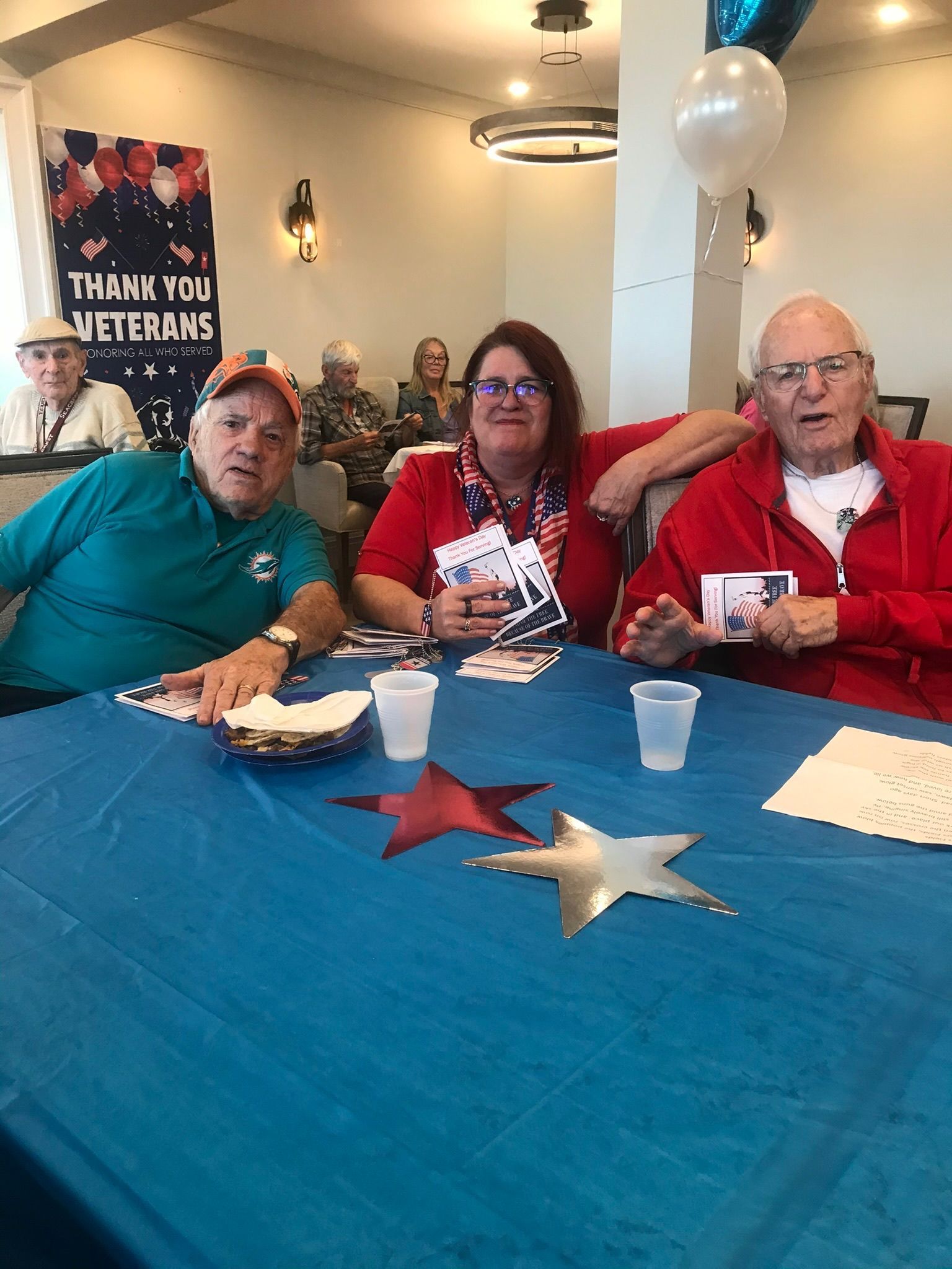 Three people at a table decorated for a veterans' event. The table has stars and blue tablecloth. People in the background.