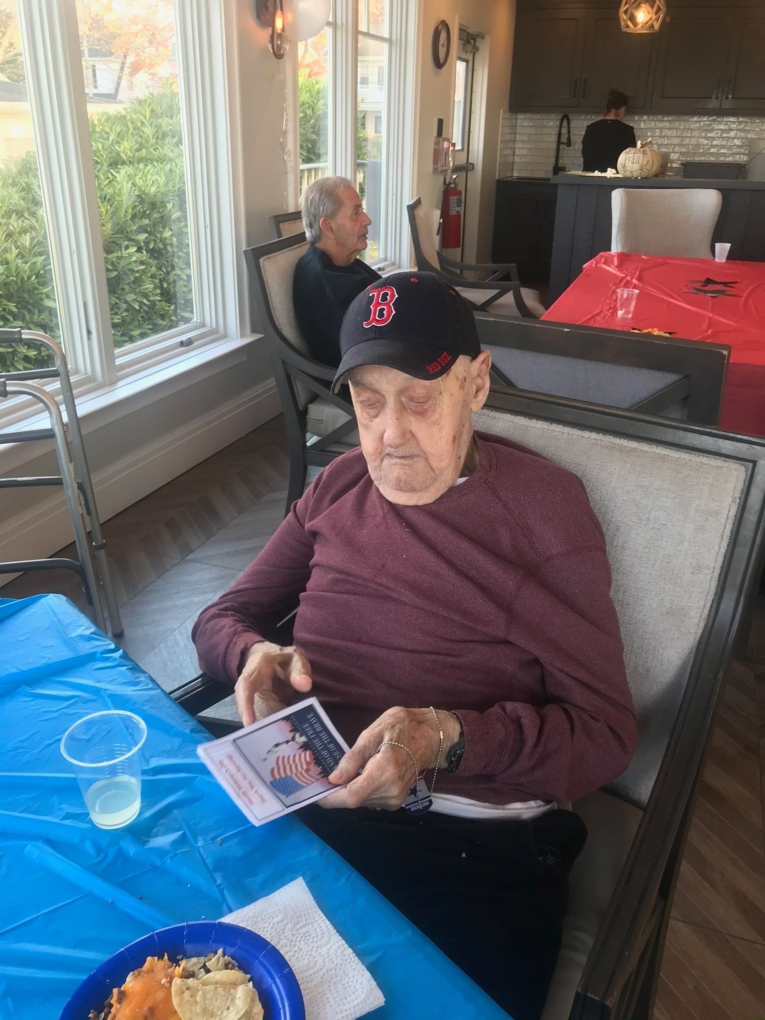 Older man wearing a baseball cap looks at a photo, seated at a table. Another person sits in the background. Bright room.