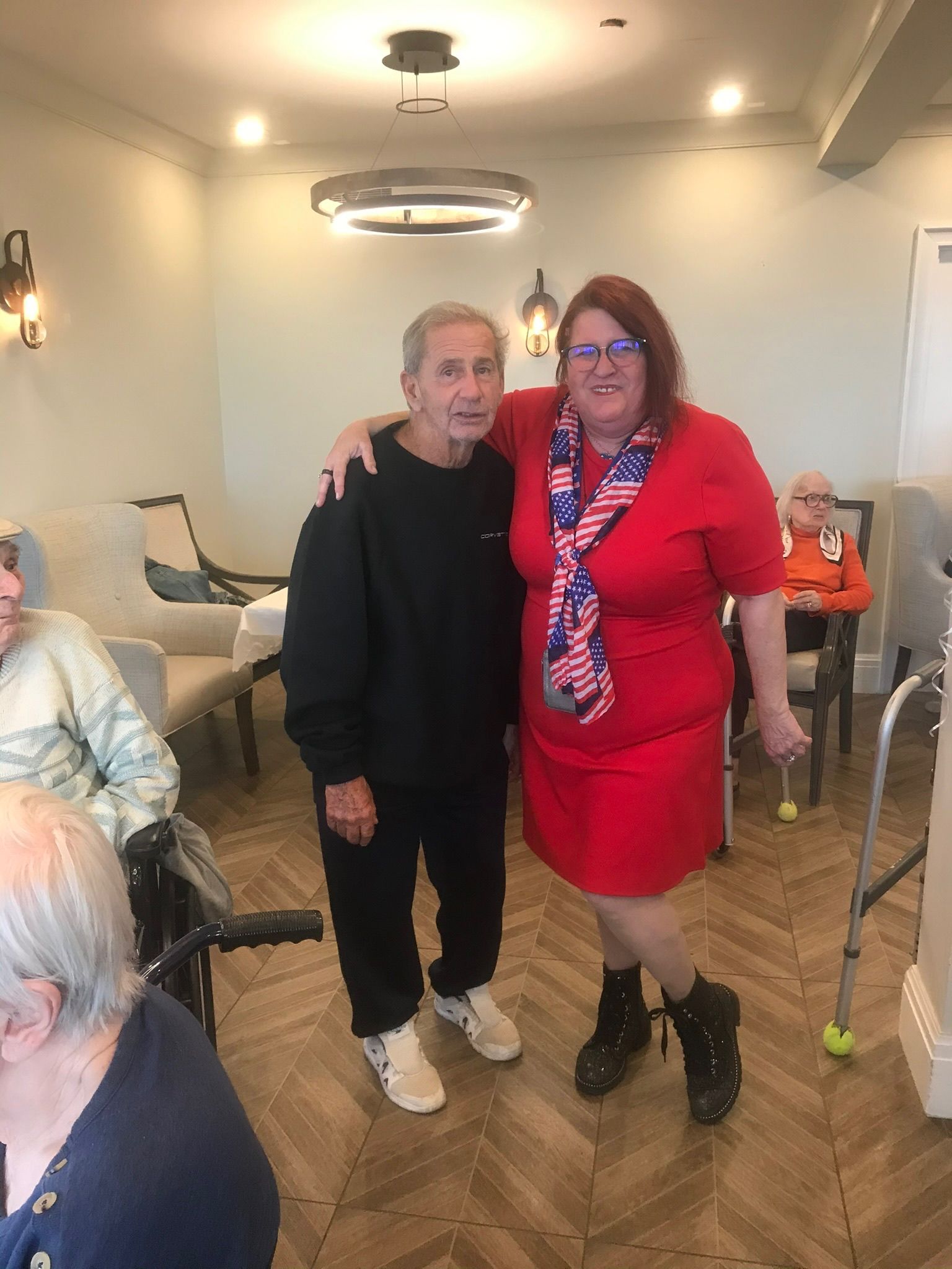 Man and woman in a room, arm around each other, woman wearing American flag scarf. Other people seated nearby.