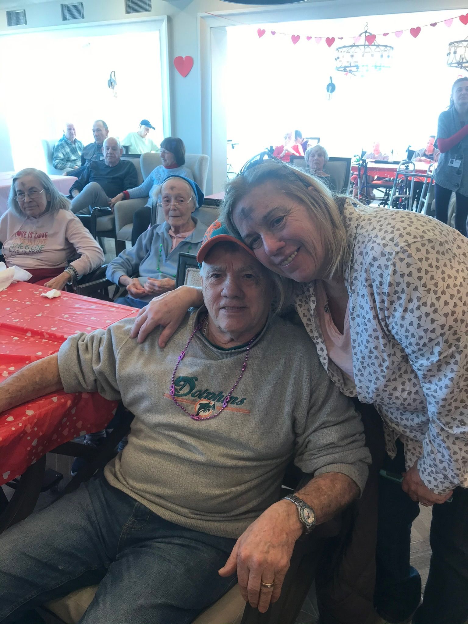Woman hugs a man in a wheelchair; they smile at the camera. In a decorated room with others.